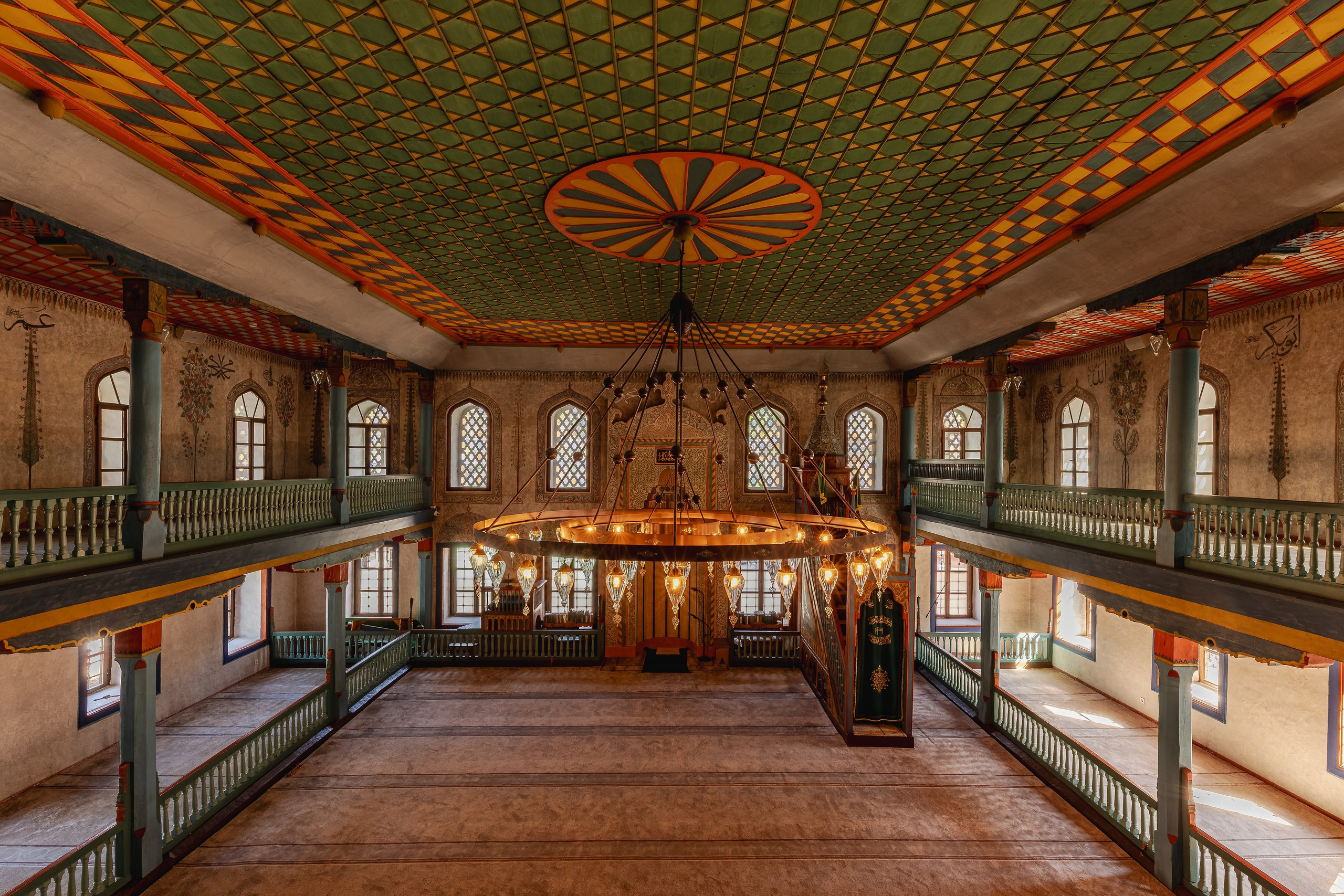 Interior architectural photography of a historic Ottoman mosque featuring geometric ceiling patterns, arched windows, wooden galleries and decorative chandeliers illuminated by natural light.