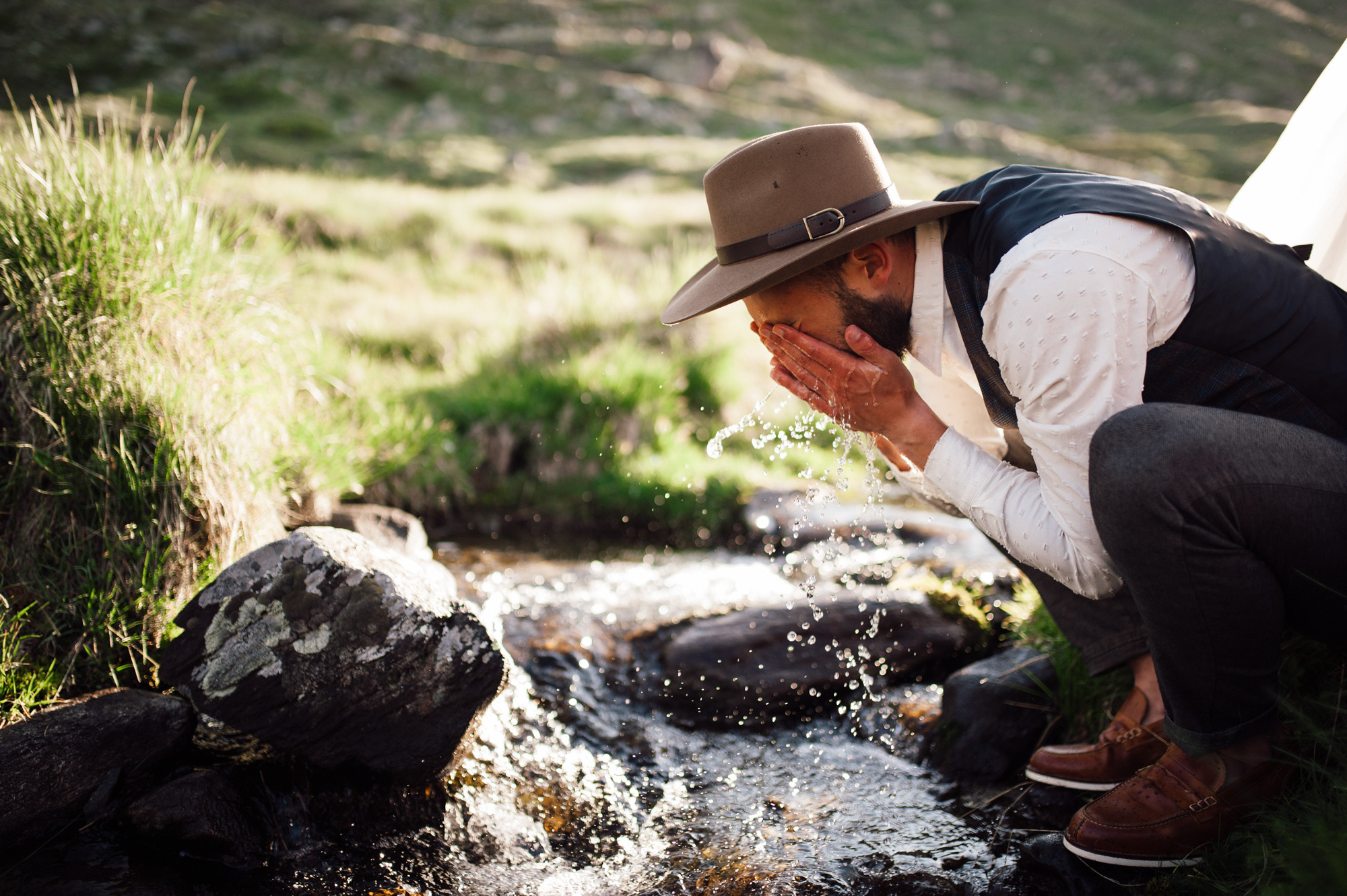 a man in a hat and vest drinking water from a stream