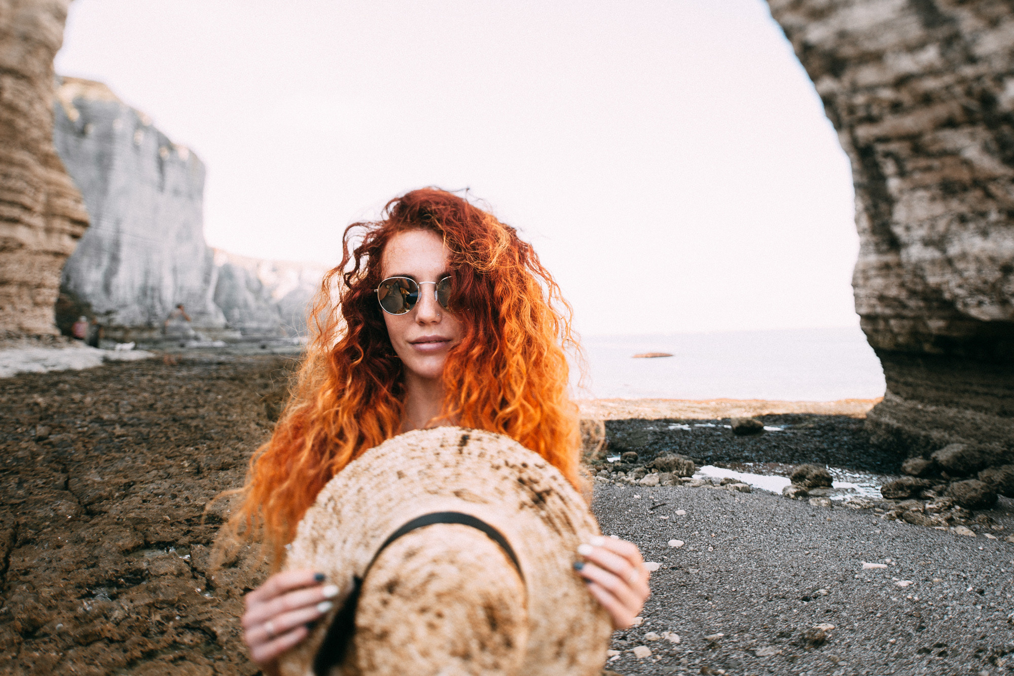 a woman with red hair and sunglasses standing on a rocky beach