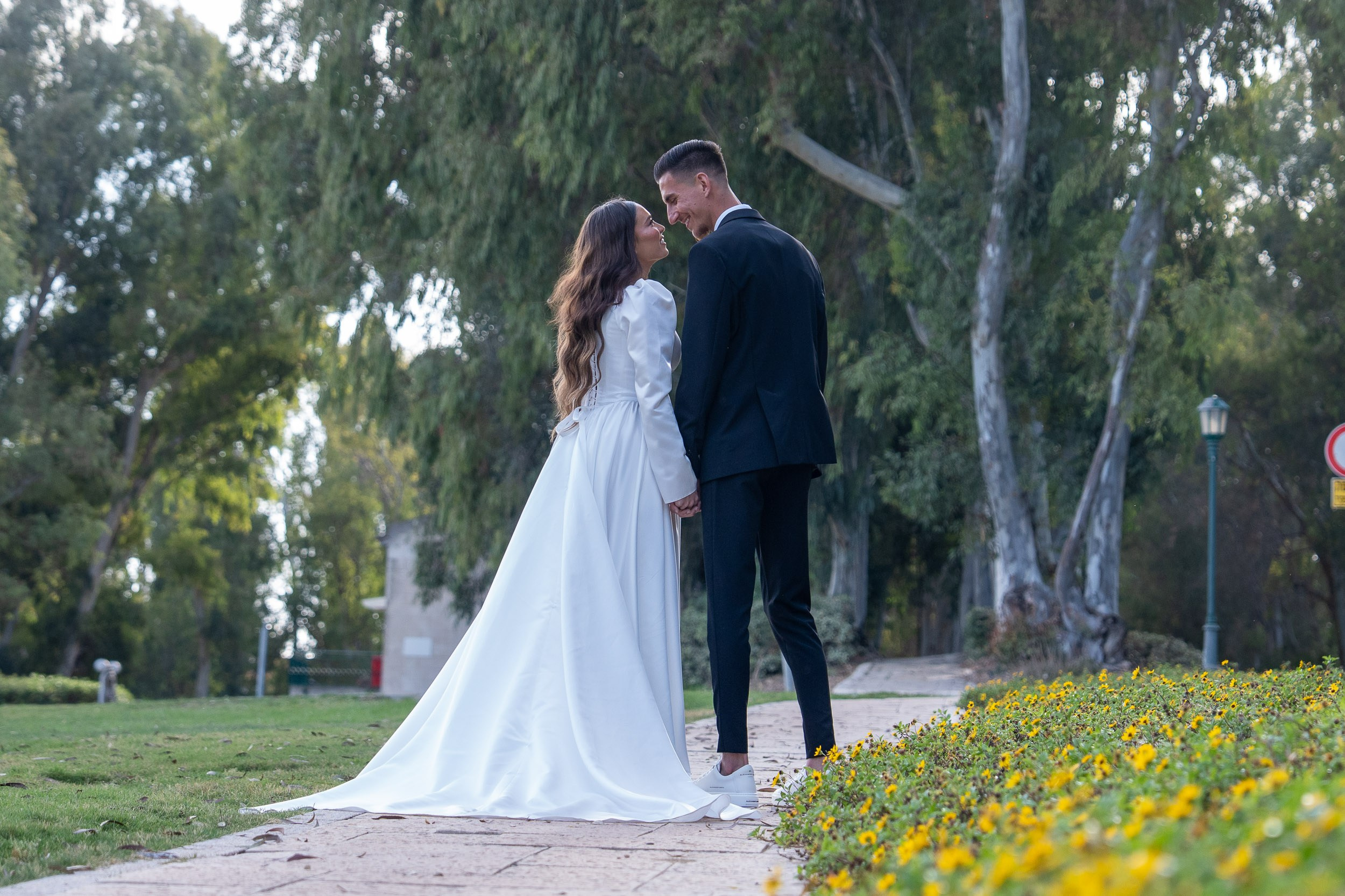 Bride Lin and groom Garik holding hands on a park pathway with yellow flowers during their photoshoot in Hadera, Israel