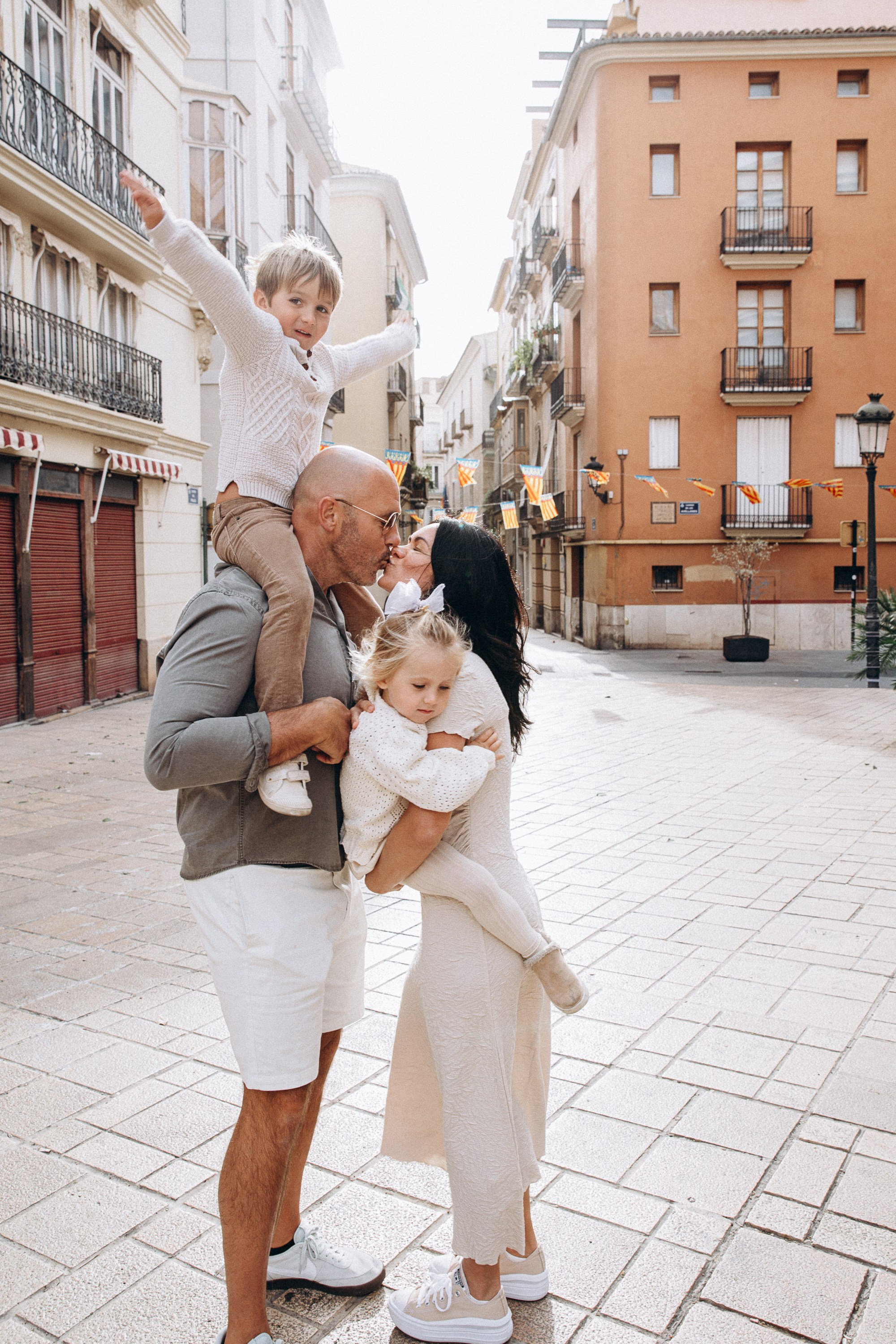 Sesión de fotos familiar en Valencia, España, con unos padres levantando alegremente a su pequeño hijo en una histórica plaza del casco antiguo rodeada de la encantadora arquitectura mediterránea. Un retrato familiar lifestyle natural que captura momentos de juego, sonrisas auténticas y la conexión familiar. Ideal para fotografía familiar, sesiones de fotos familiares, retratos infantiles, fotografía de vacaciones y fotografía familiar de destino en Valencia y toda España.