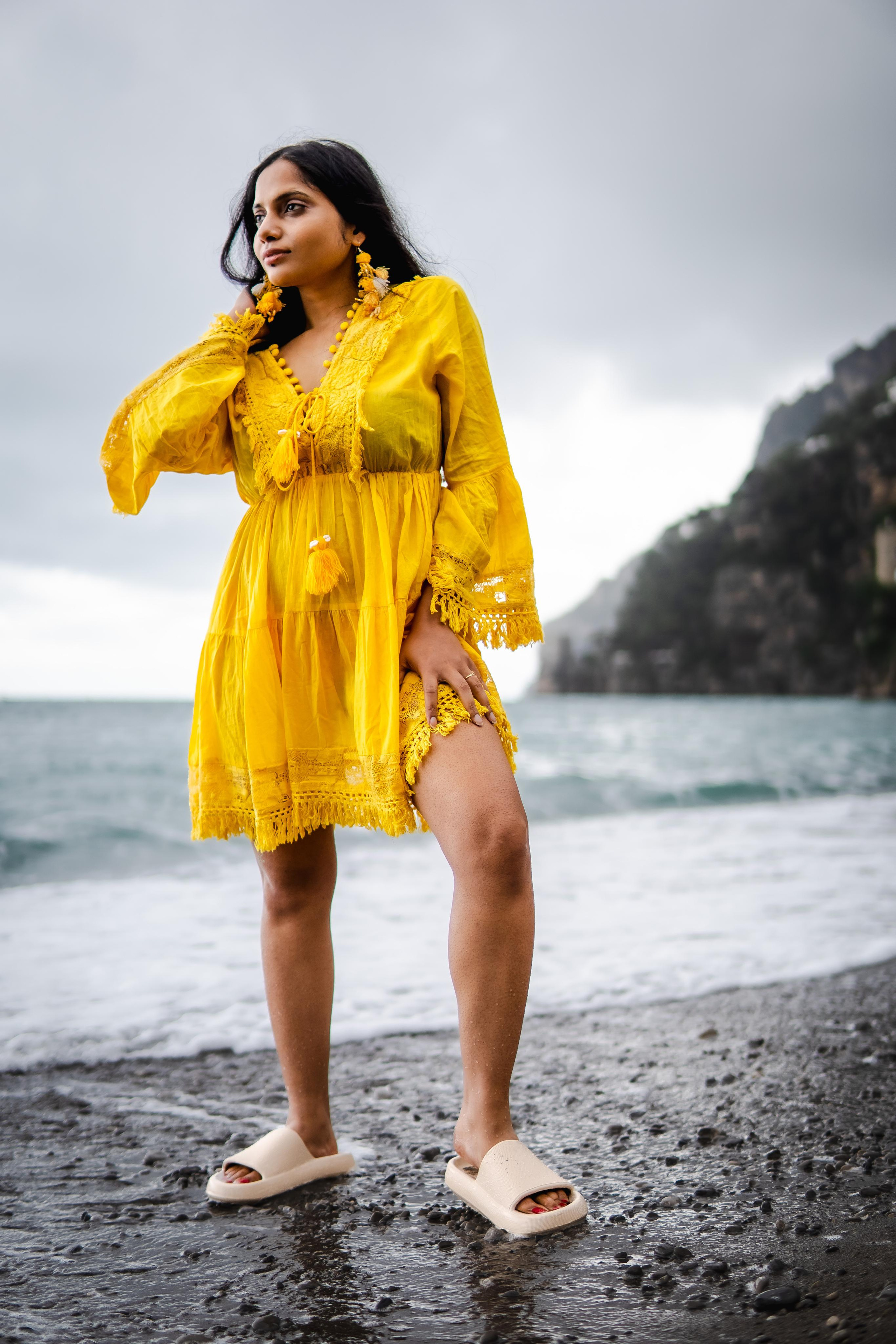 a woman wearing a yellow beach dress standing near a beach in positano and playing with her hair