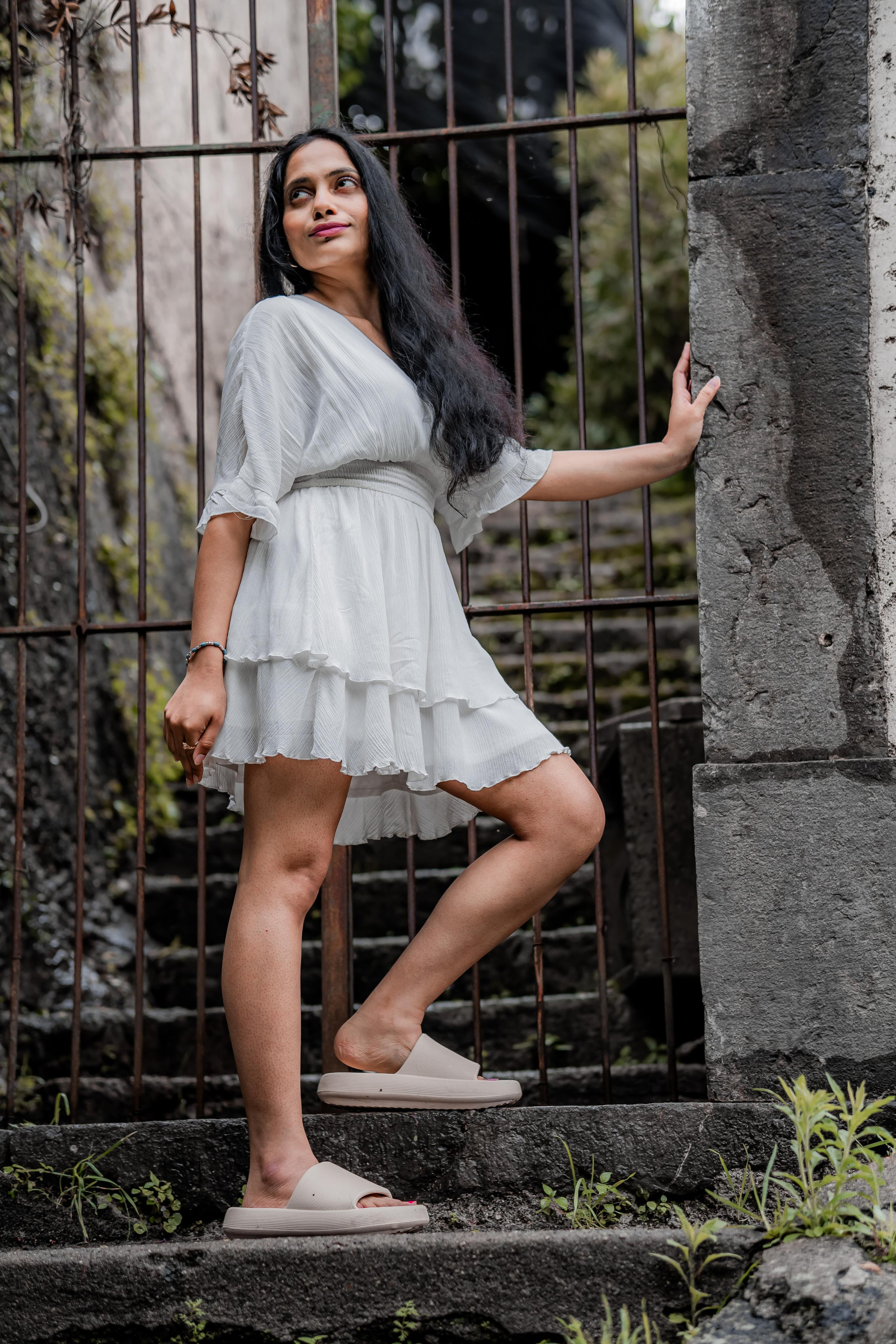 a woman standing near a vintage old gate in a hilly area wearing white mini frill dress and looking at a distance