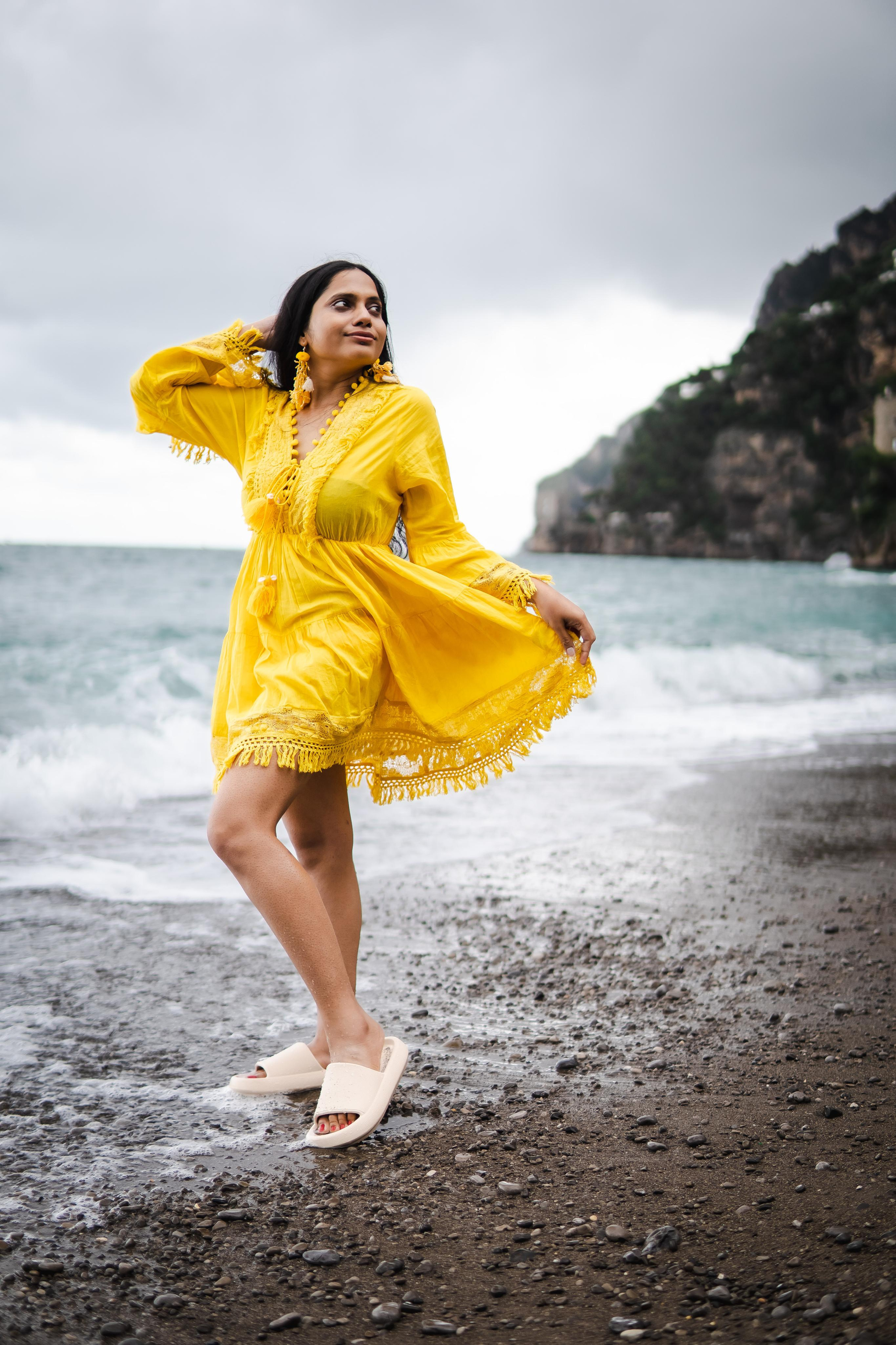 a woman wearing a yellow beach dress standing near a beach in positano and playing with her hair