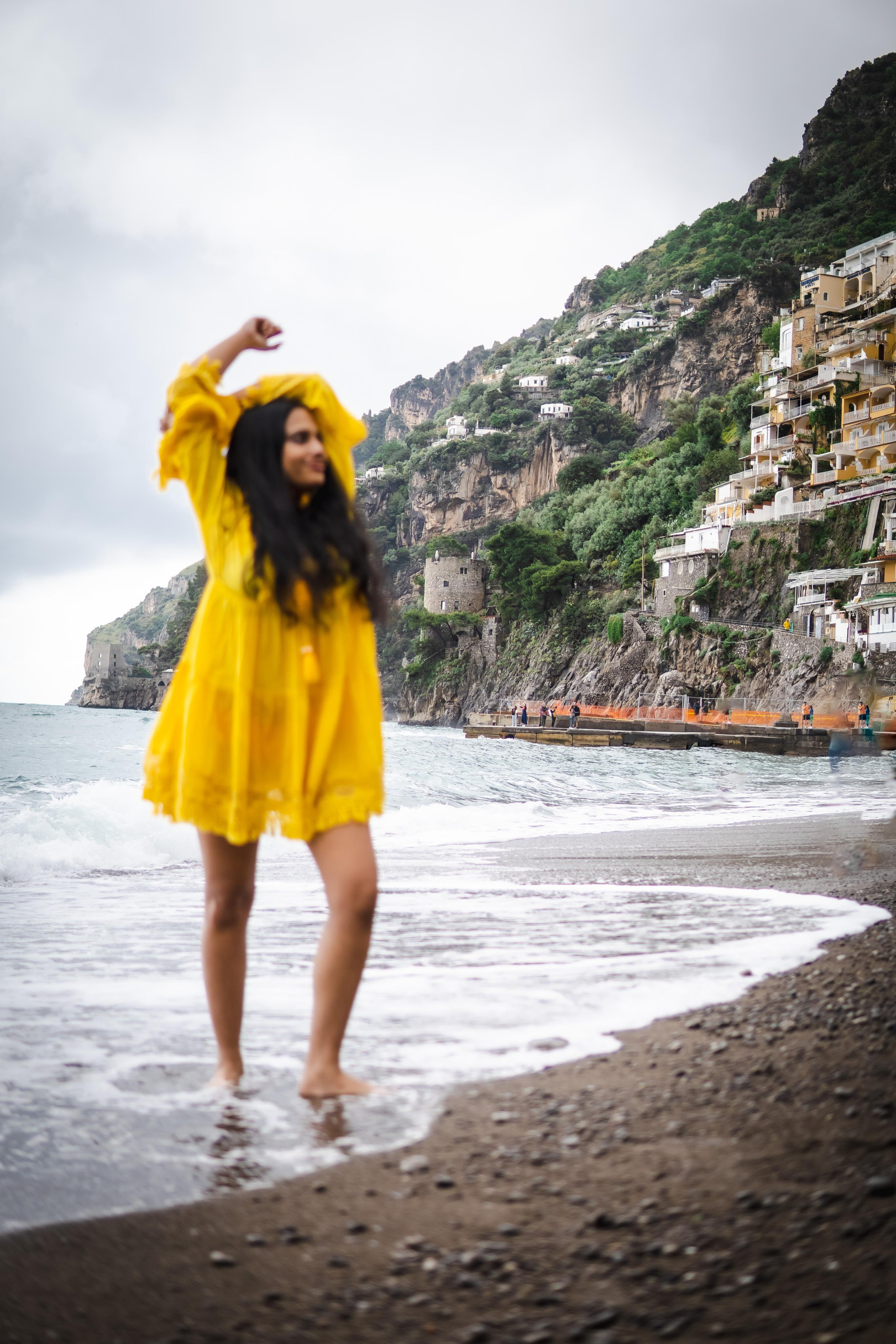 a woman wearing a yellow beach dress and standing near a beach in positano with her arms raised 