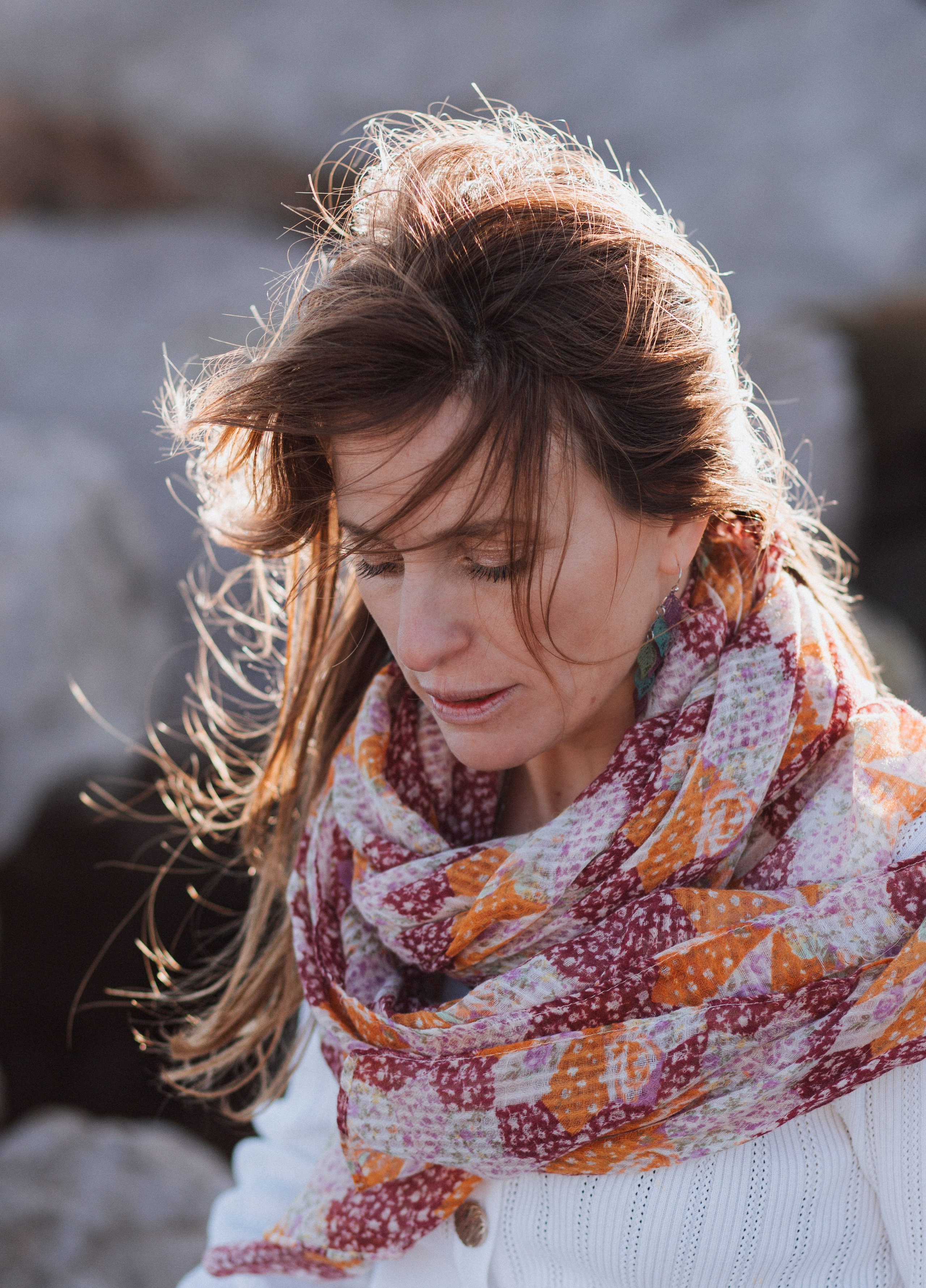 Ветреный портрет женщины в шарфе с узором и белом свитере, смотрящей вниз, с солнечным светом на лице у скалистого побережья в Валенсии, Испания