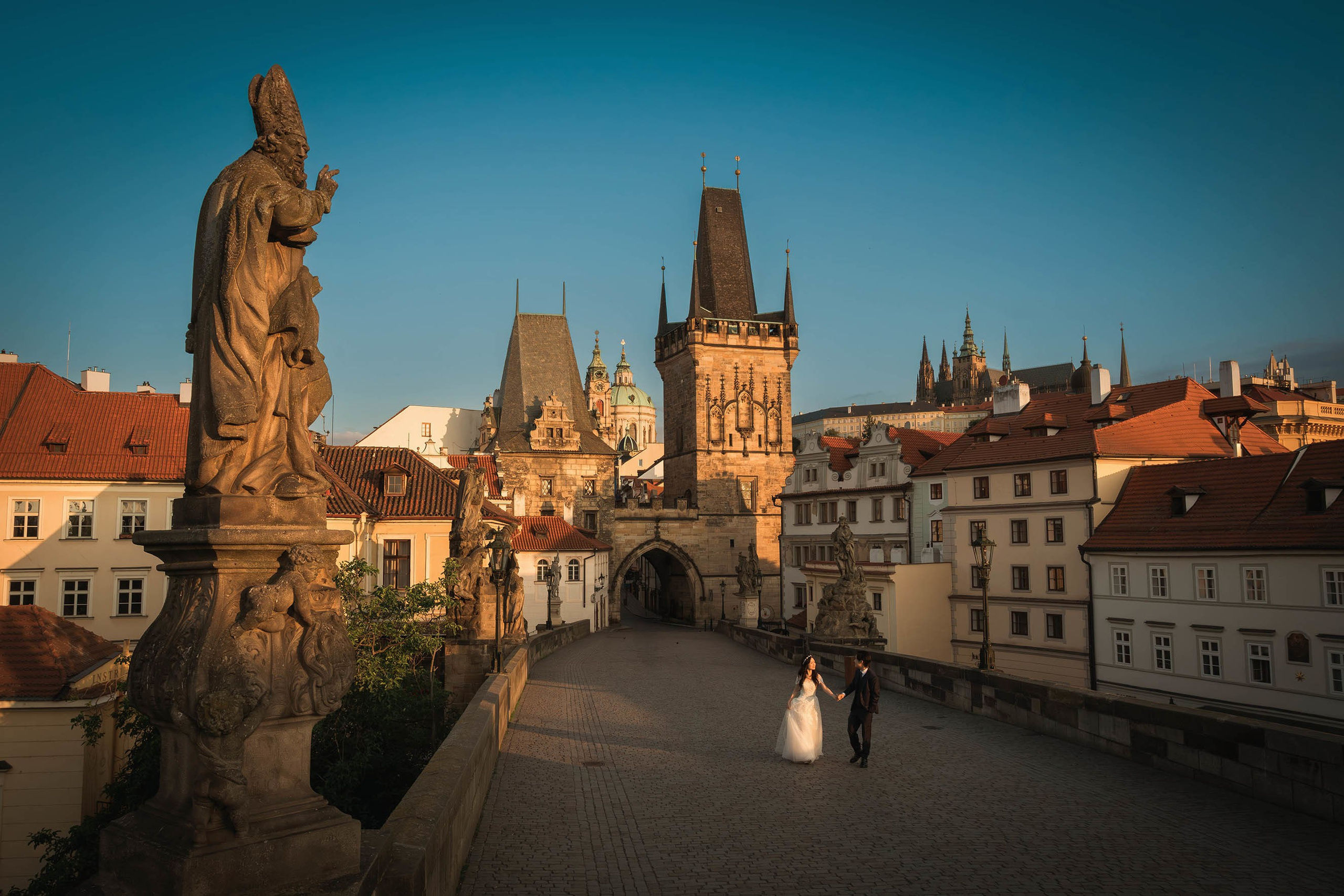 As a statue looms high above, down below a smiling bride and groom walk all alone atop the Charles Bridge at sunrise as Prague Castle is seen in the background