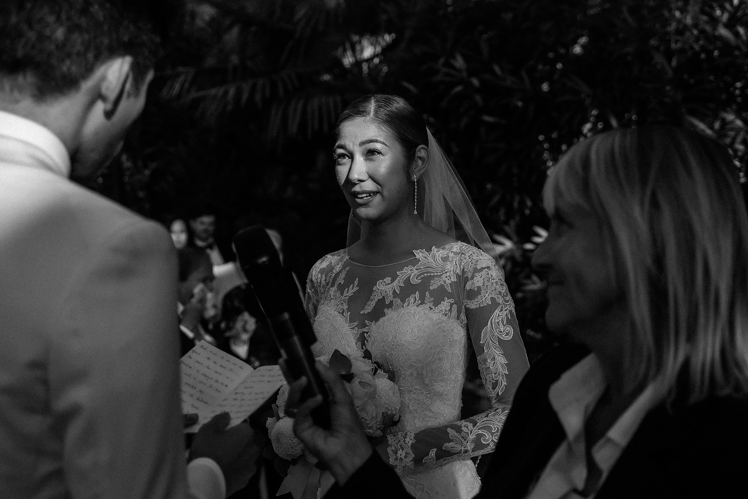 Bride gazes at the groom emotionally as he speaks during the ceremony, holding her bouquet.