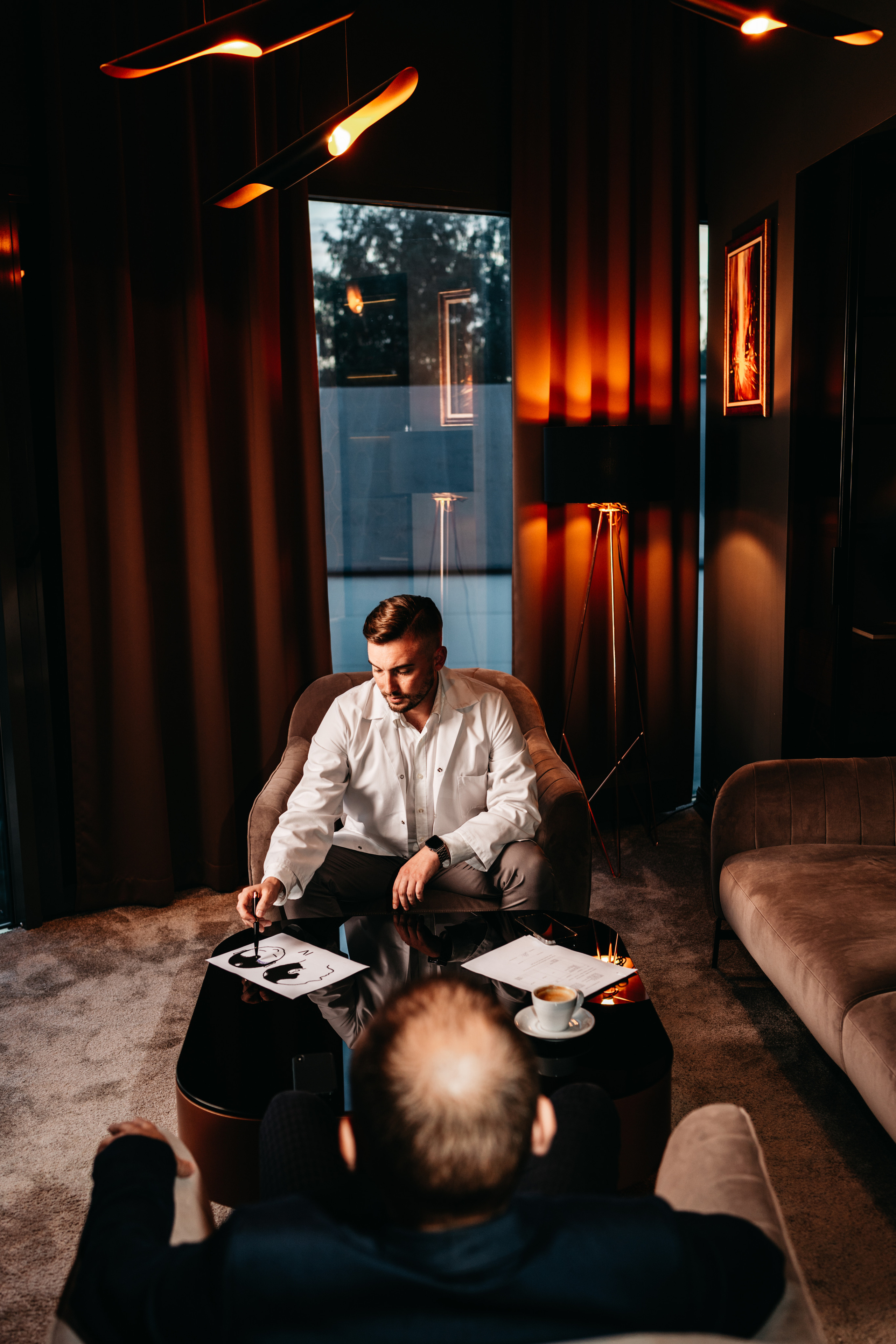 A man sitting in a stylish, dimly lit interior, dressed in casual attire, exuding sophistication.