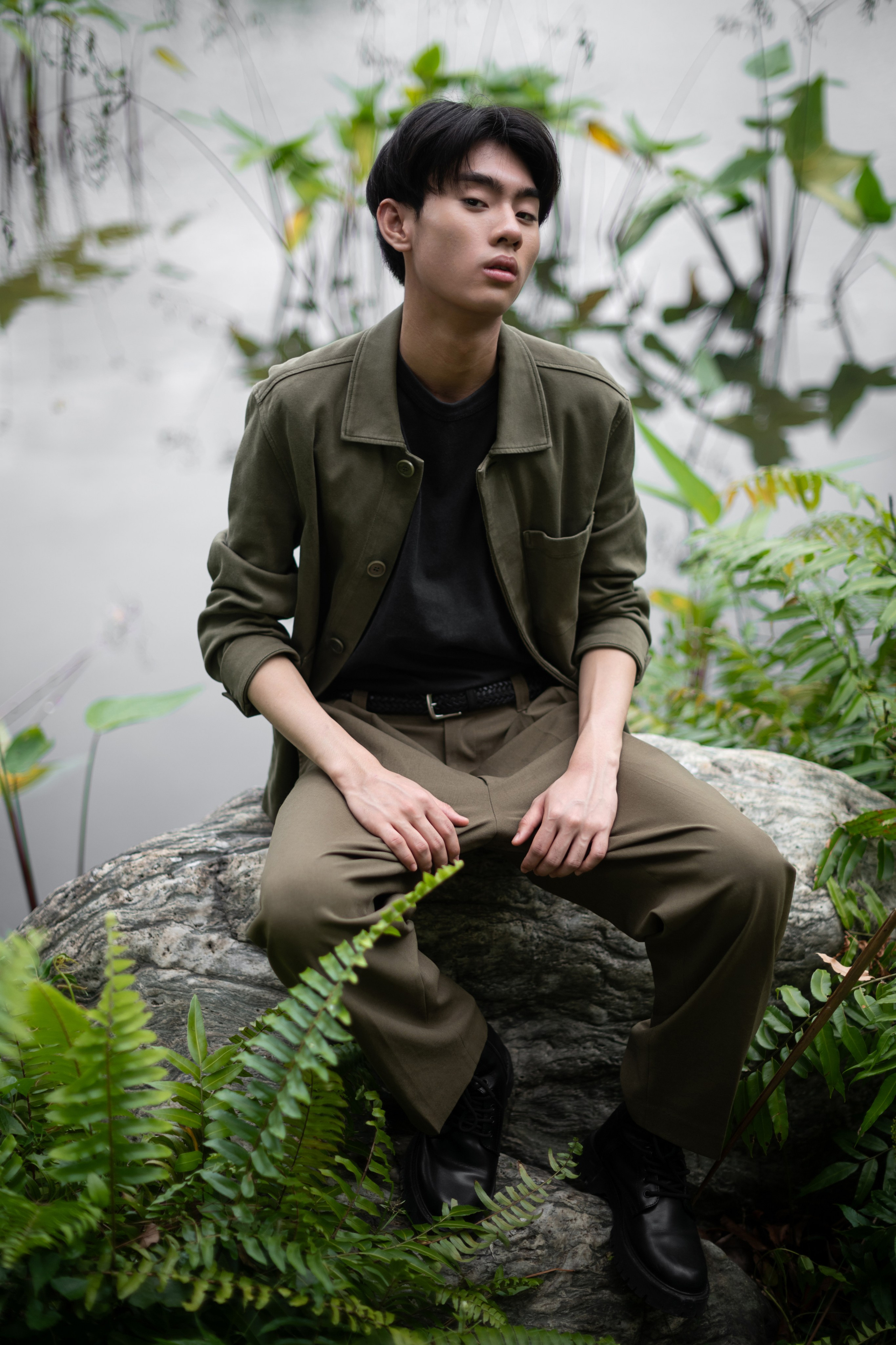 Young Asian man in olive jacket and trousers sitting on a rock, hands on knees, surrounded by ferns with a blurred lotus pond behind him.