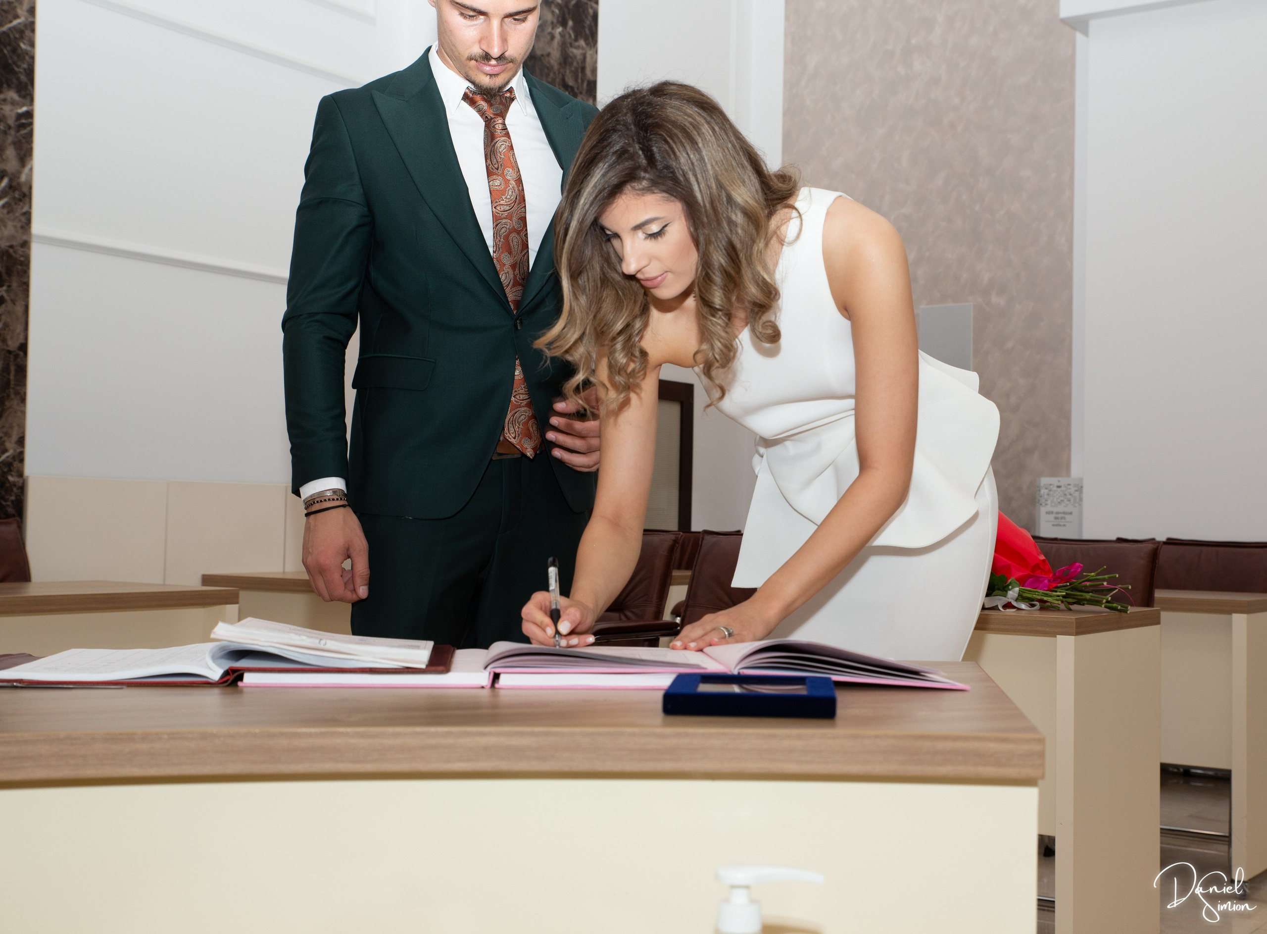 Bride in white dress signing a document as groom stands next to her in a suit.