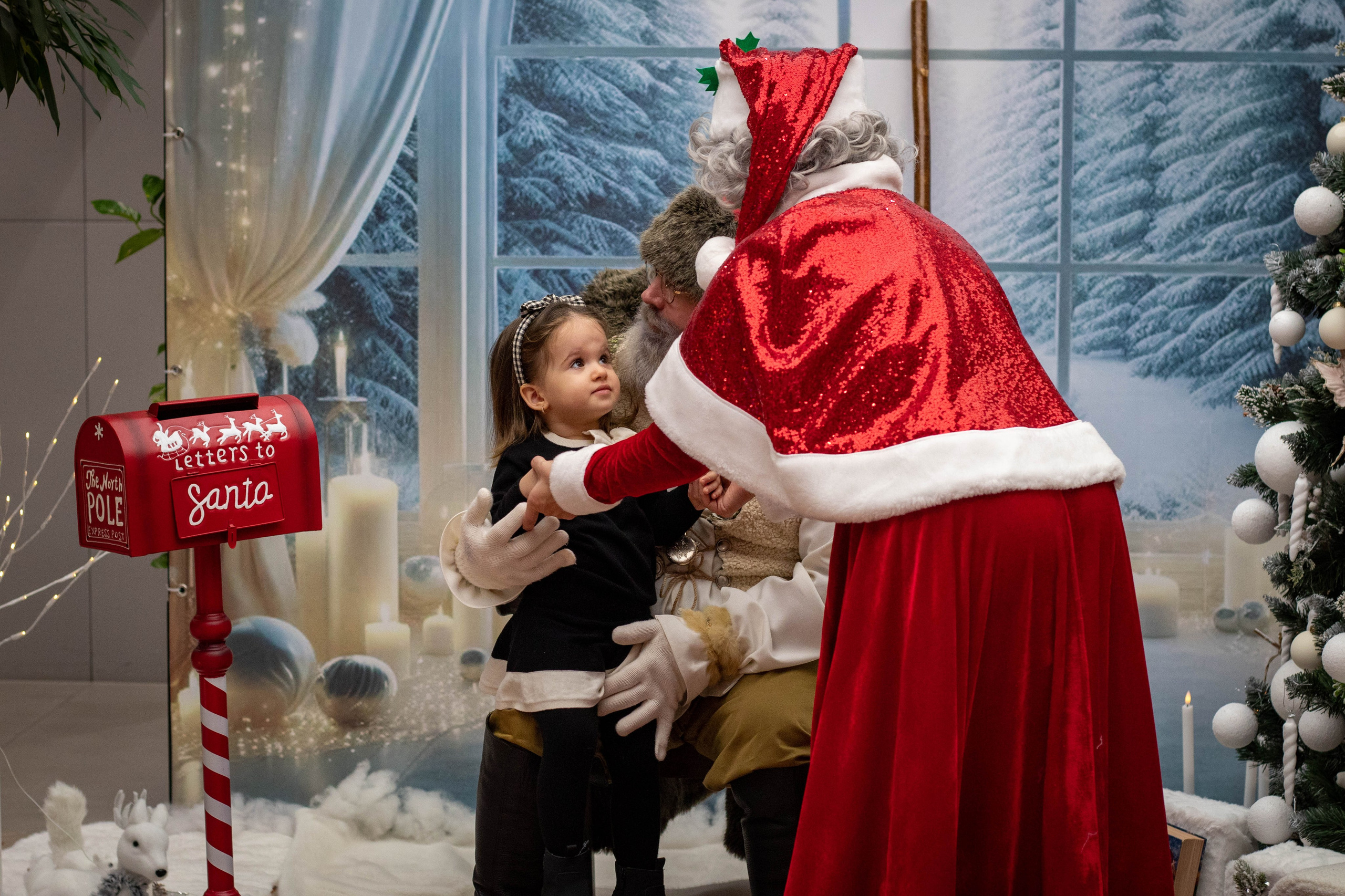 Santa Claus sitting near a decorated tree, handing a gift to a young child during a holiday celebration.