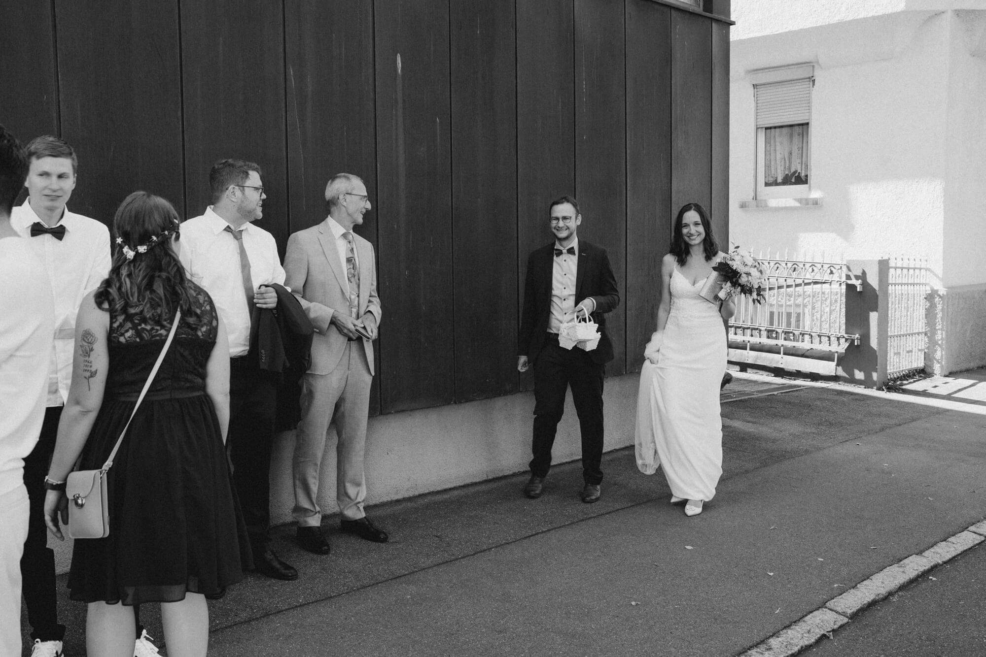 Bride arriving on foot with wedding guests outside the ceremony venue in Stuttgart, bouquet in hand; black and white