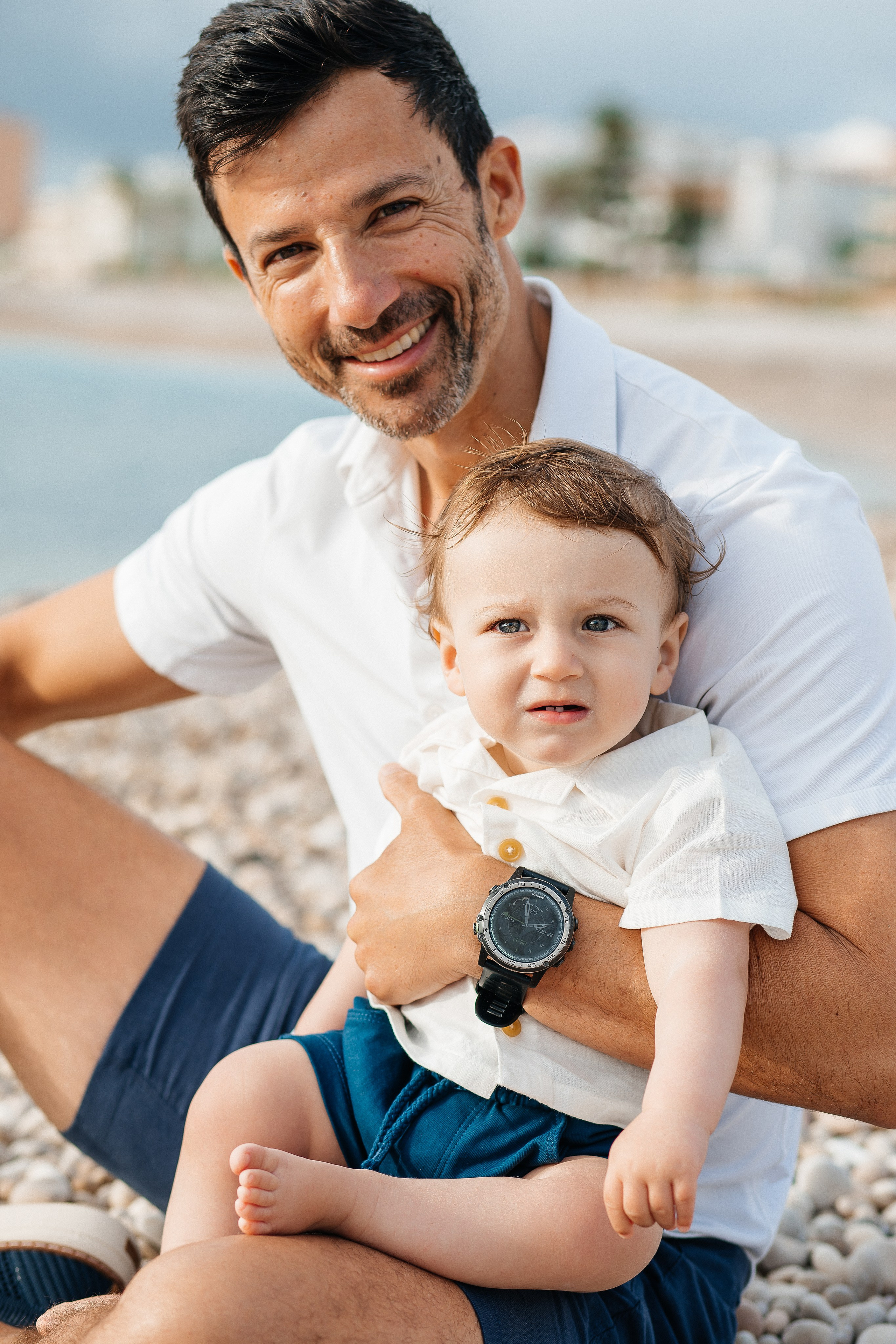 Sesión de fotos familiar en Valencia, España, con un padre sonriente sosteniendo a su pequeña hija en una playa del Mediterráneo durante la hora dorada. Un retrato lifestyle natural de padre e hija que captura felicidad, amor y momentos familiares inolvidables. Ideal para fotografía familiar, fotografía de padre e hija, sesiones familiares, retratos infantiles, fotografía de playa y fotografía familiar de destino en Valencia y toda España.