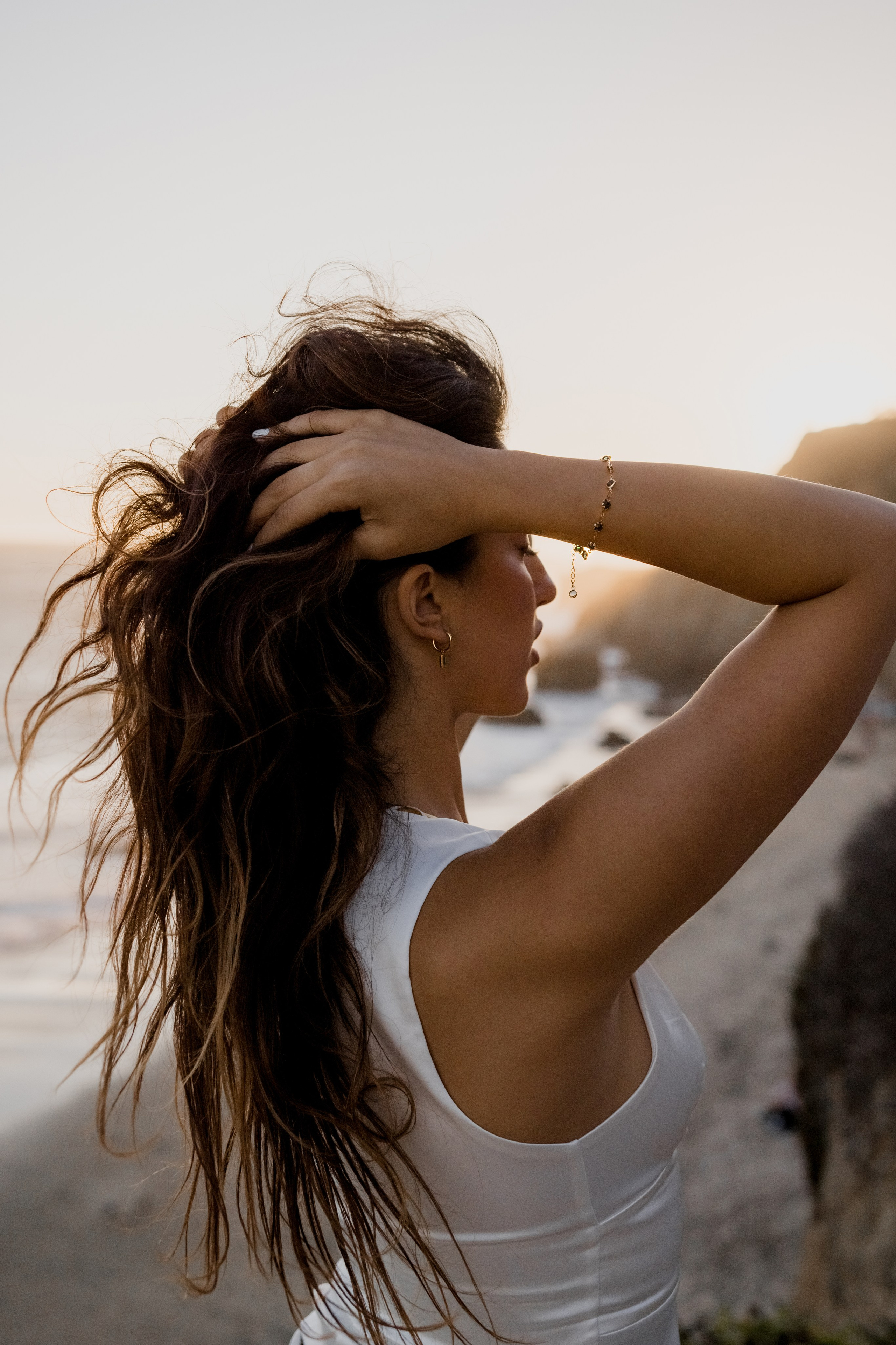 Fashion model styled in a white dress, posing on the beach with flowing hair.