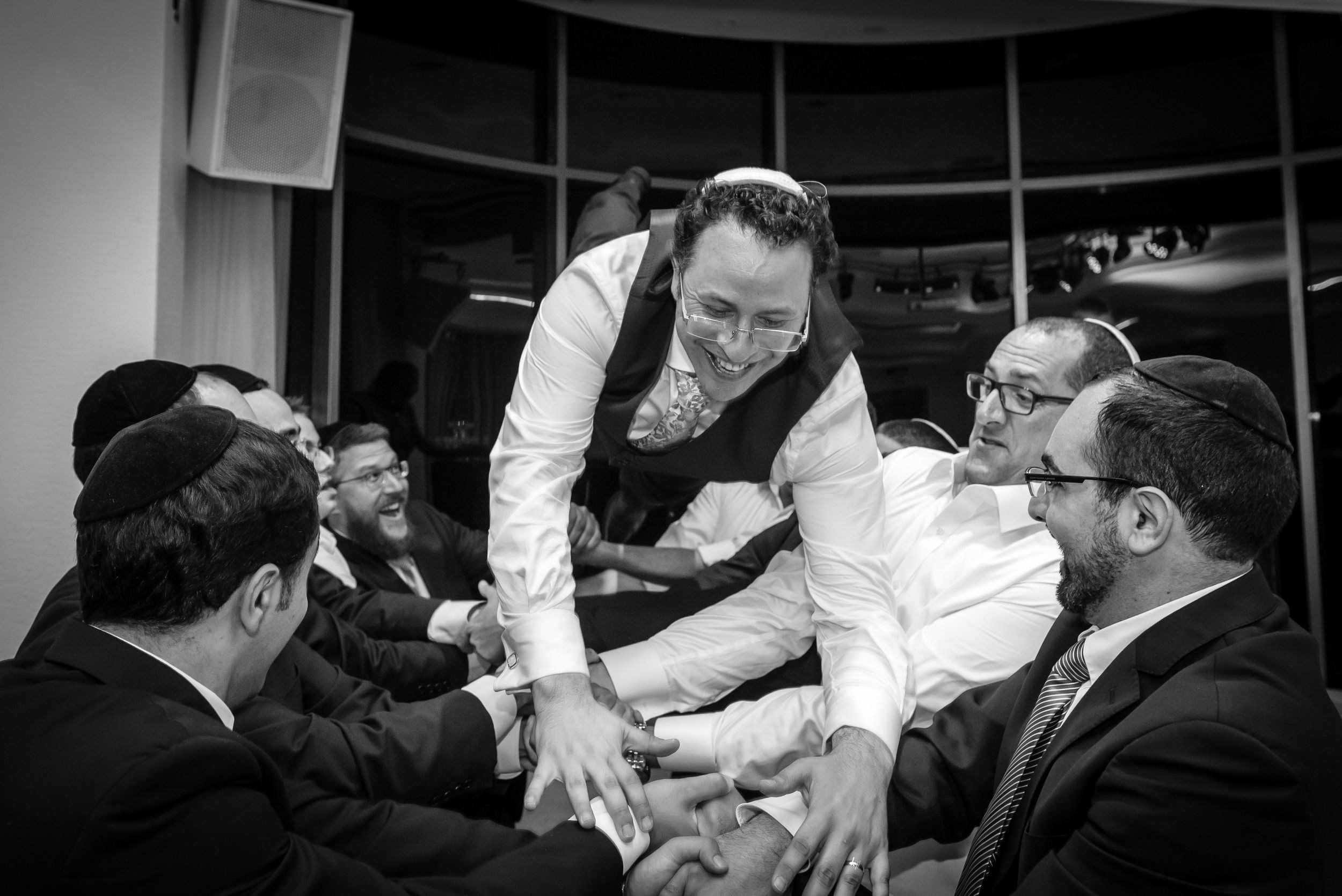 A dynamic black and white capture of pure joy. The groom is lifted by friends in a lively wedding celebration, surrounded by laughter and energy.