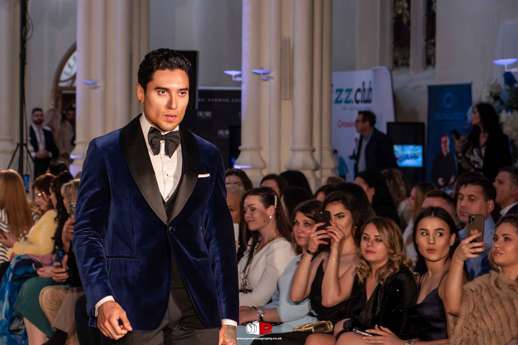  A male model in a stylish blue velvet tuxedo is walking down the runway at a glamorous fashion event with an attentive audience.