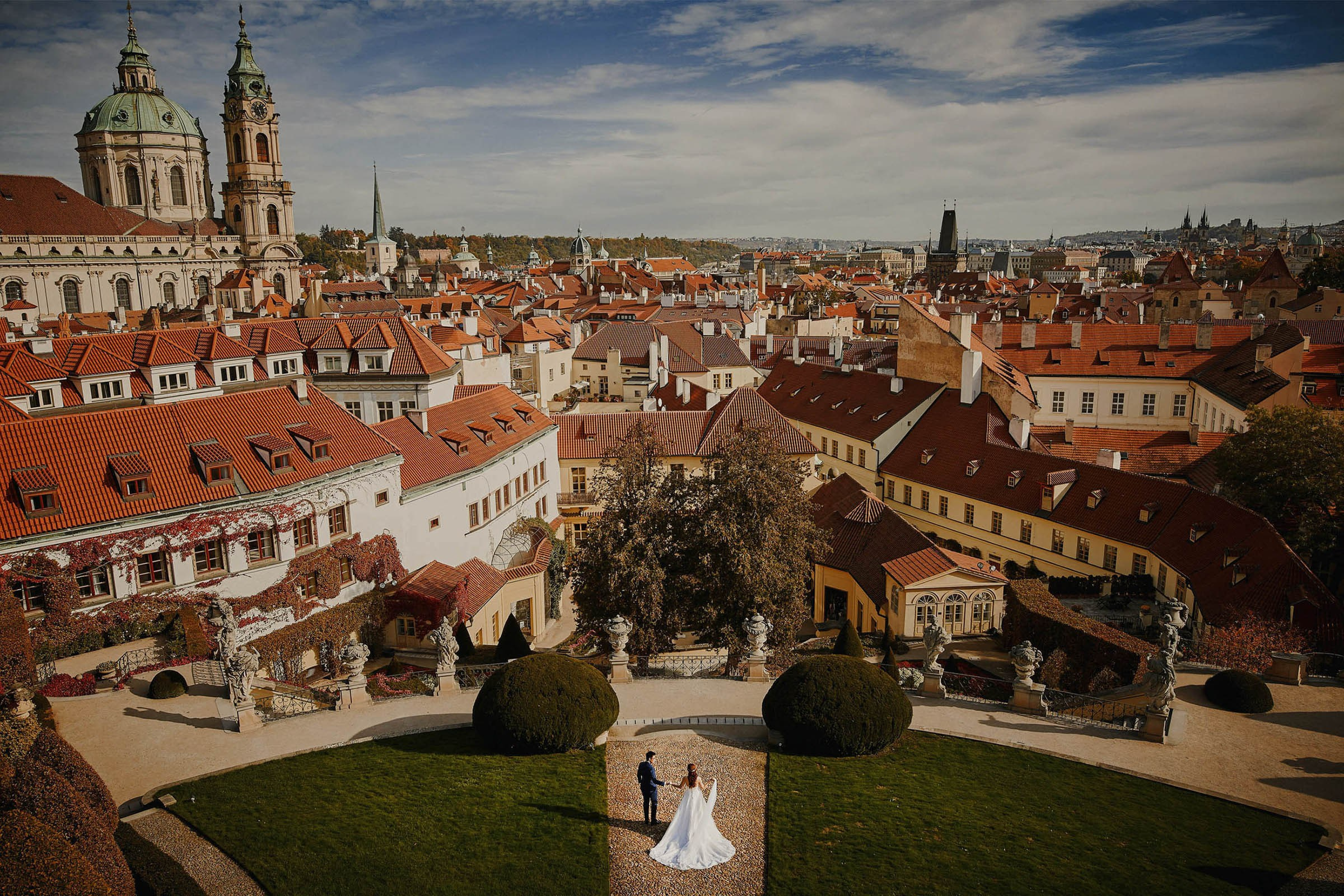 A view from above the historic Baroque Vrtba Garden showing the Prague skyline with St. Nicholas Church (l) and Charles Bridge (r) as a bride & groom walk hand in hand below.