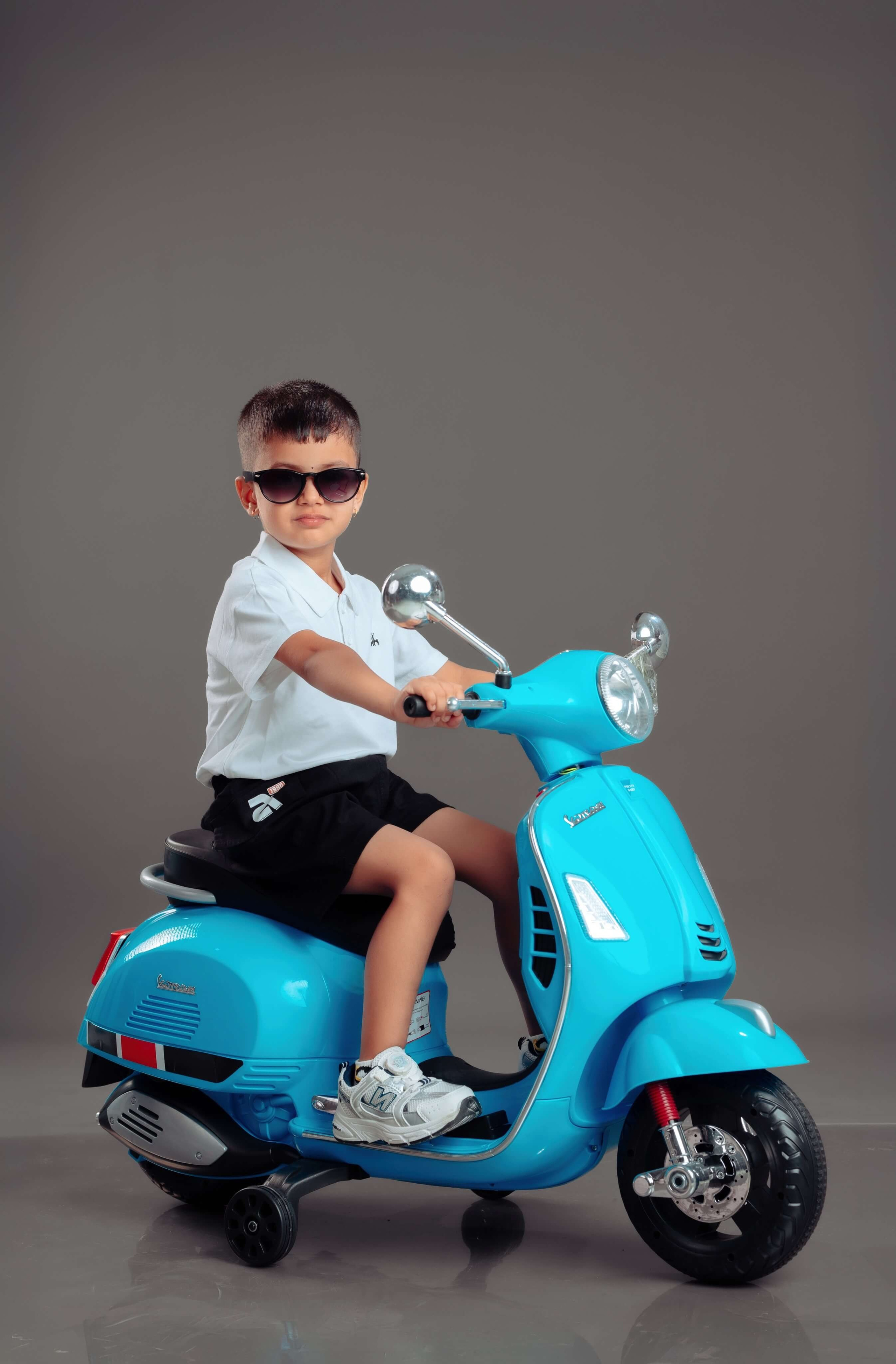 A stylish baby boy wearing sunglasses and riding a bright blue toy scooter in a studio shoot