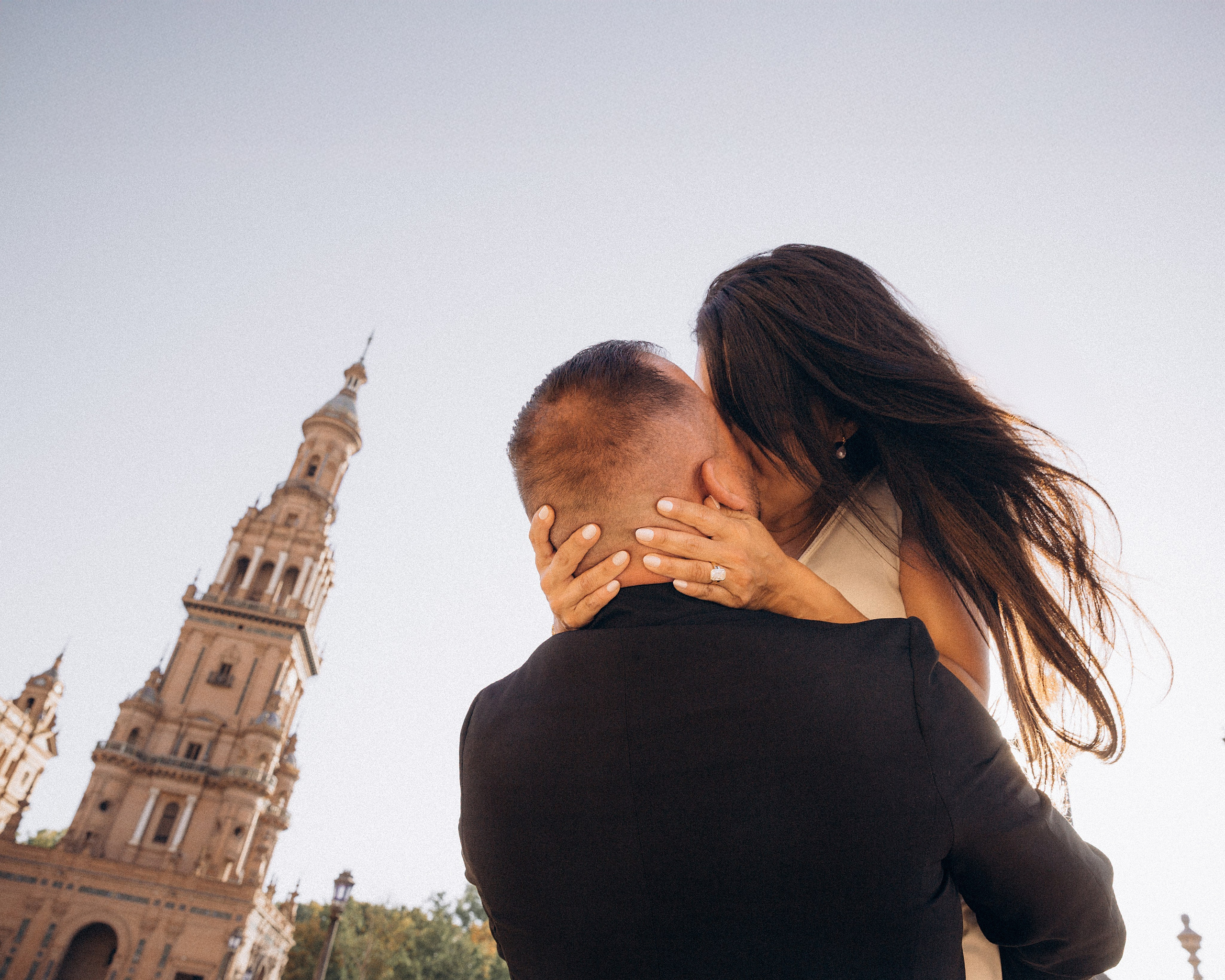 Close-up of the couple kissing with the Plaza de España tower rising behind them in Sevilla. Romantic sunset portrait from their intimate elopement-style wedding in Andalusia.