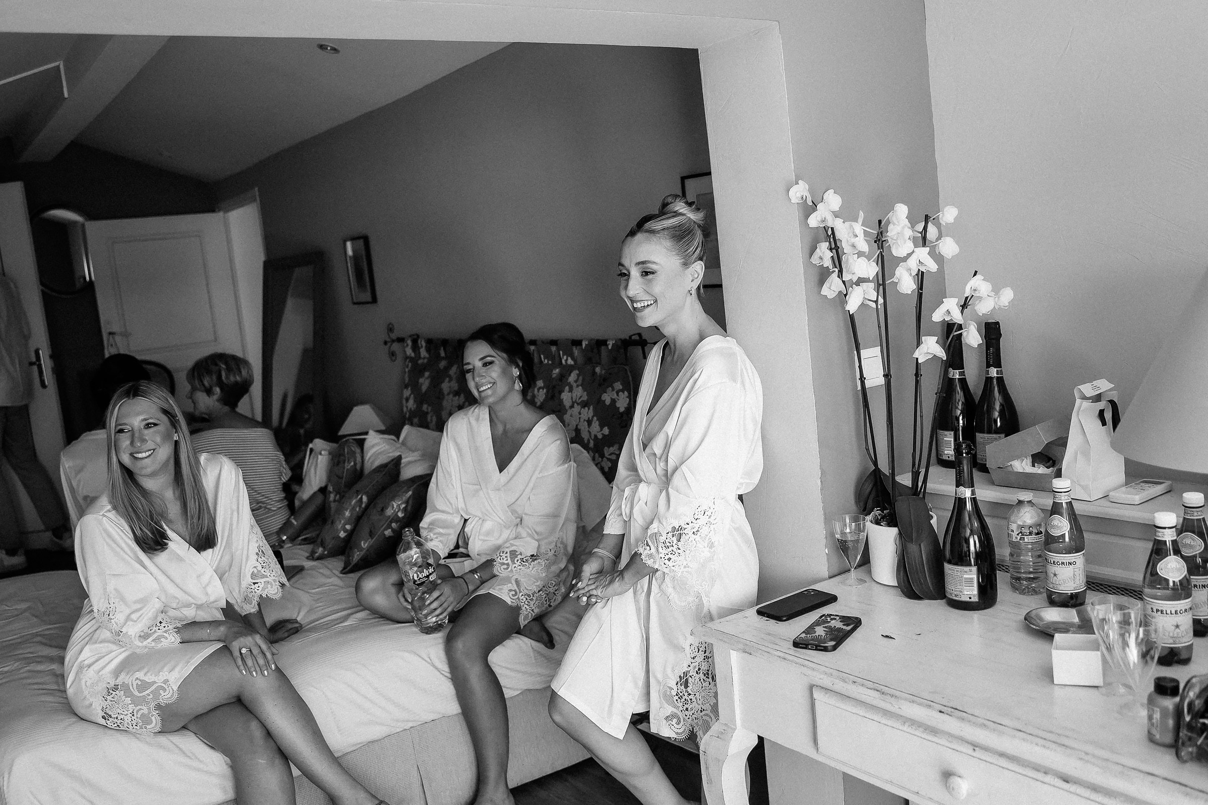 A candid moment of bridesmaids sharing laughter and stories in their white robes in a softly lit room with a feminine and elegant ambiance.