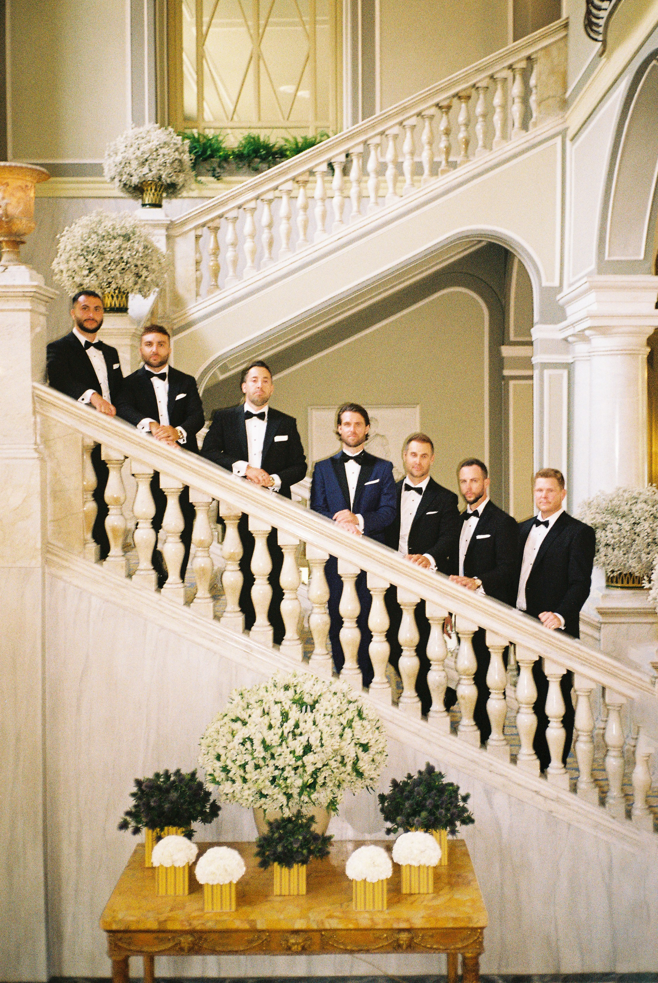 Groom and groomsmen in tuxedos on grand staircase, surrounded by white floral arrangements in an elegant hall.