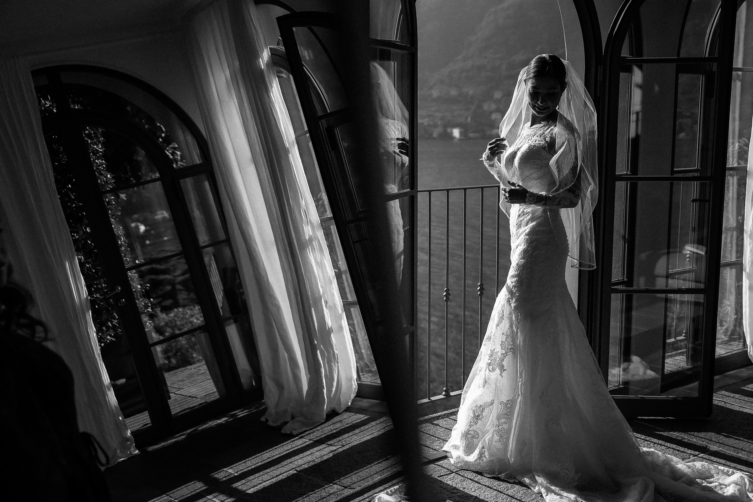 Bride stands by open balcony doors, her veil illuminated, overlooking Lake Como.
