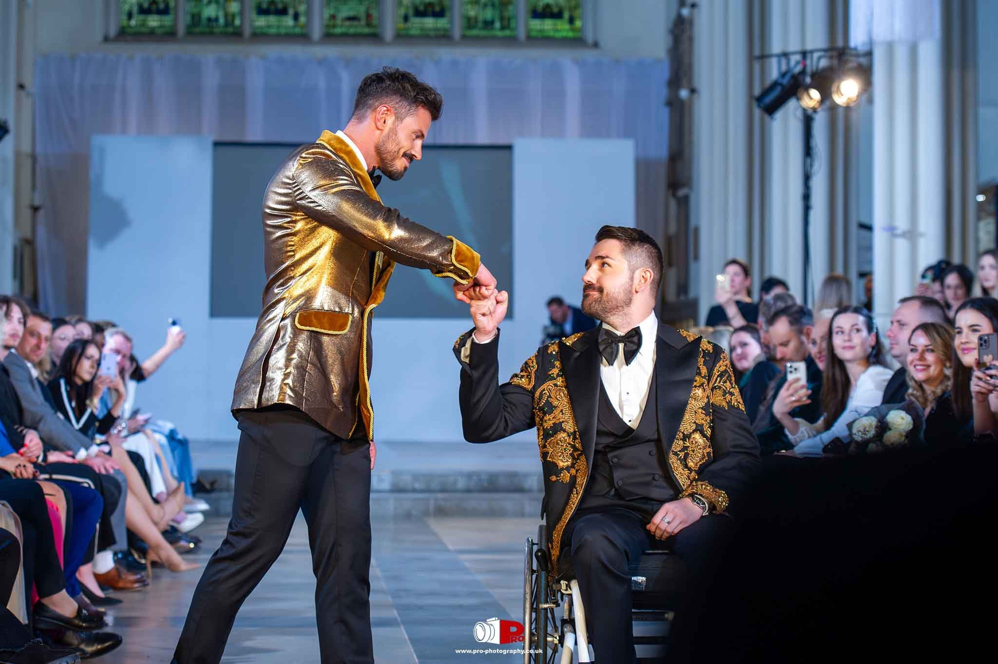 A wheelchair user and a standing model greet each other on the runway, showcasing inclusive fashion at a stylish event.