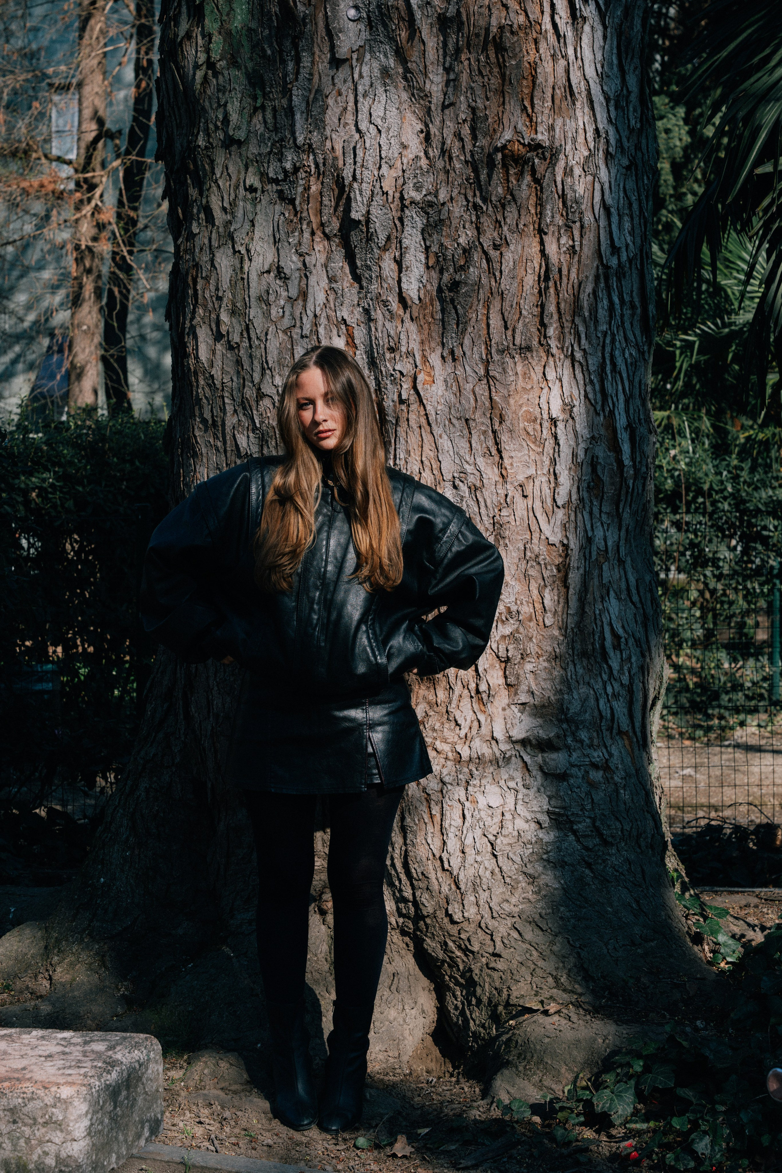женская фотосессия в парке Милан девушка в объемной кожаной куртке стоит в тени дерева 