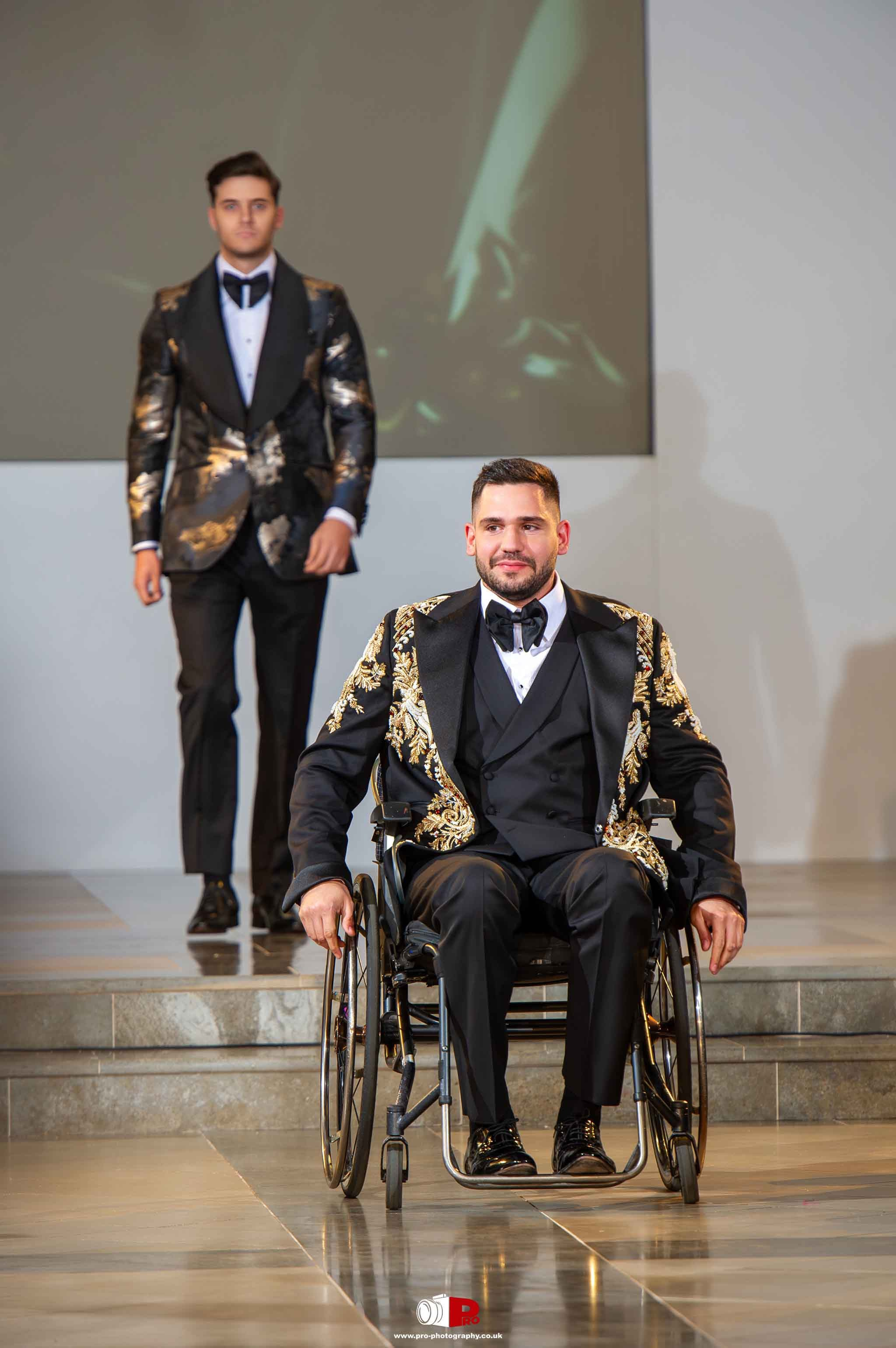 A male model in a wheelchair confidently walks the runway in an elegant black and gold embroidered suit, with another male model following behind him.