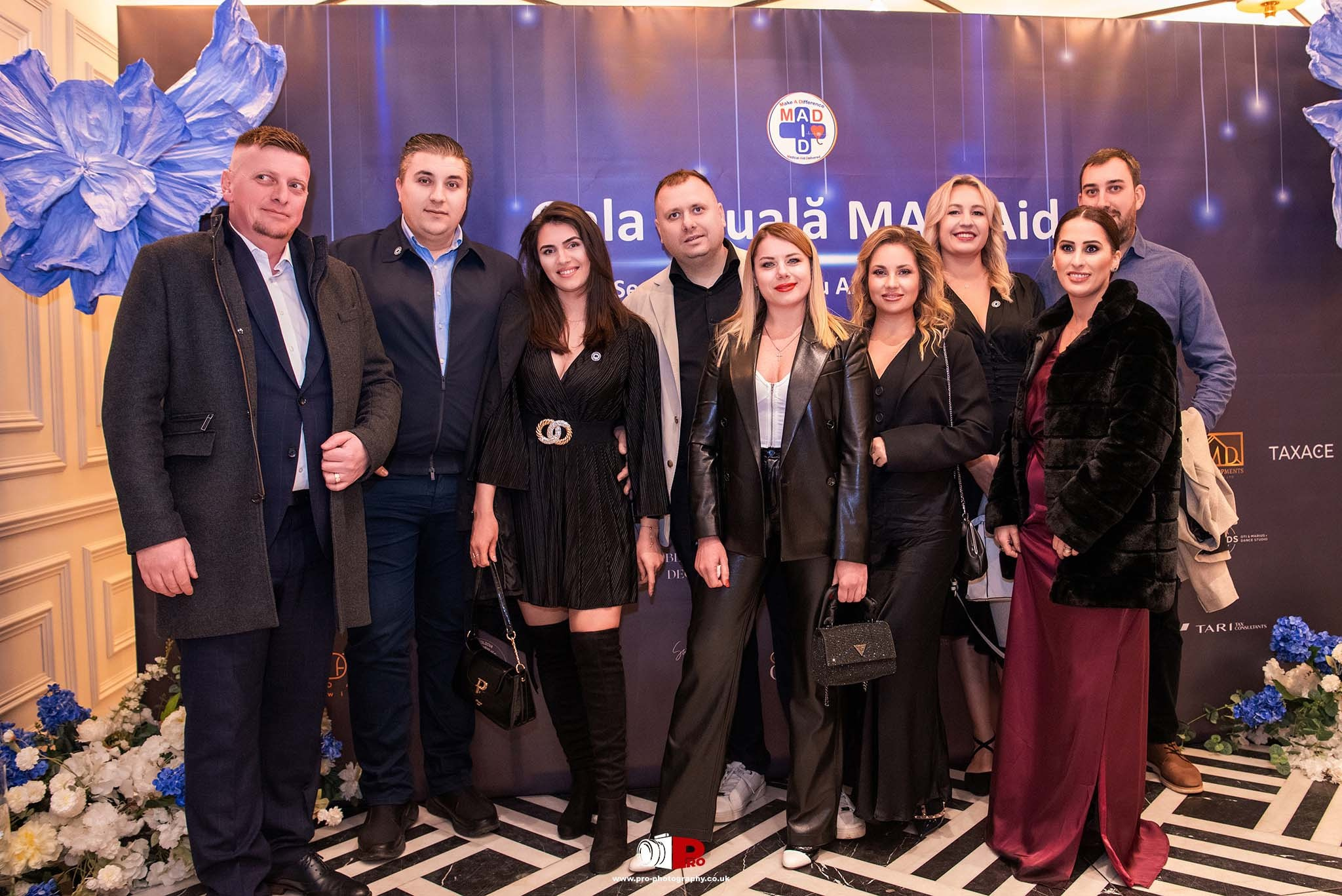A group of elegantly dressed attendees posing at a gala celebration with a decorated backdrop featuring blue flowers.