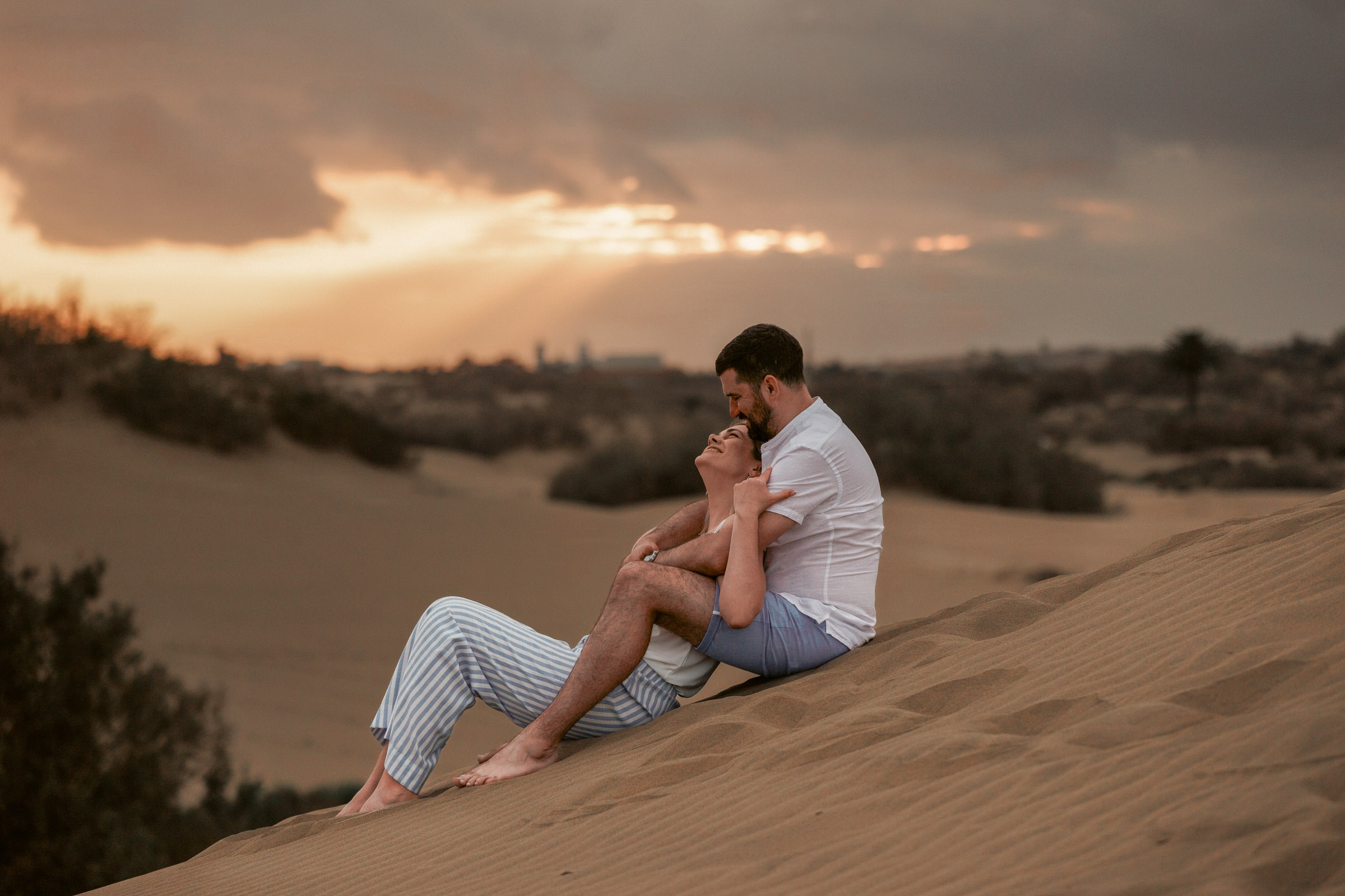 Couple Photographer Gran Canaria Maspalomas - A couple sits closely together on a sand dune, watching a sunset with rays of light piercing through cloudy skies.