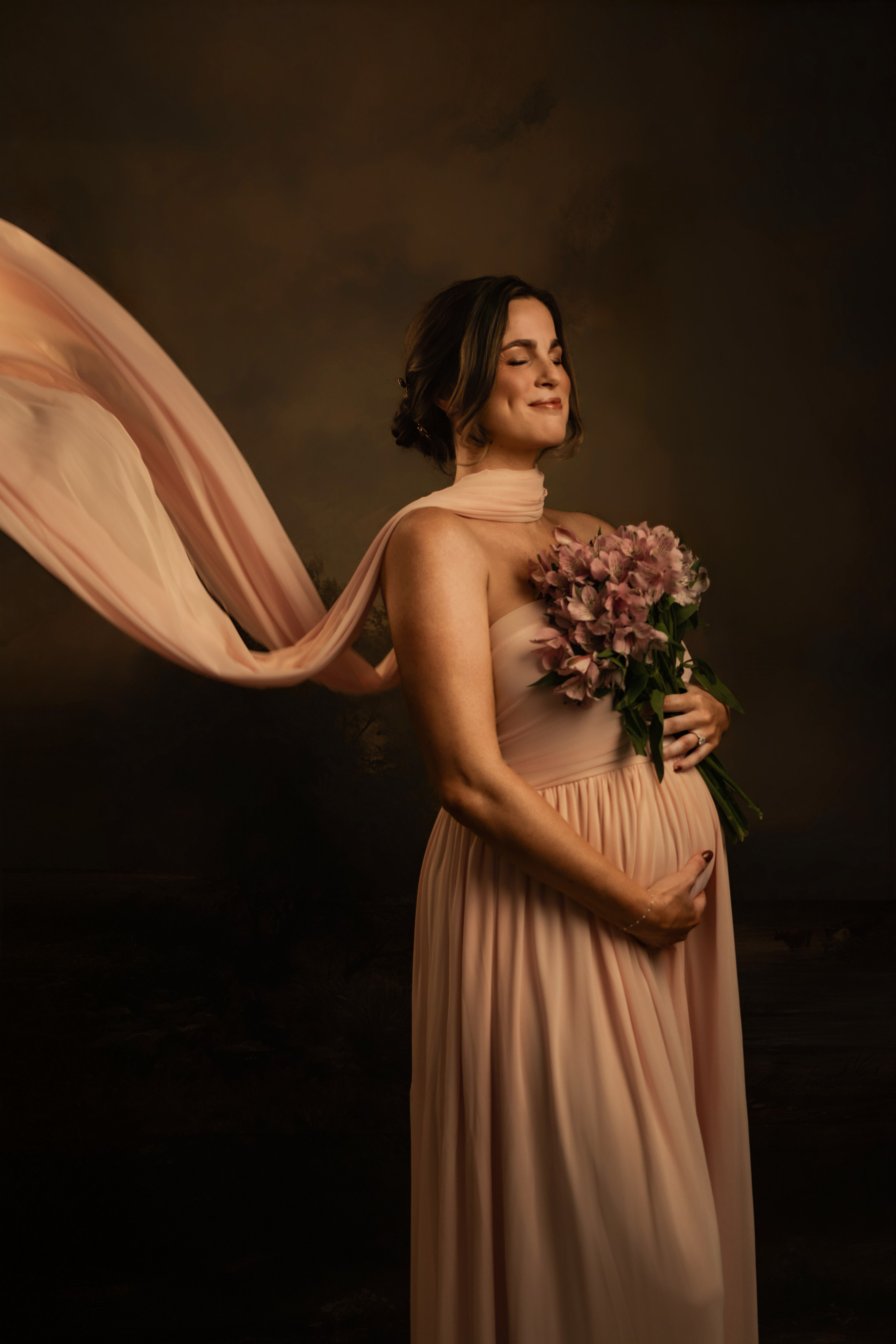 Pregnant woman in a blush gown with fabric flying behind her, studio