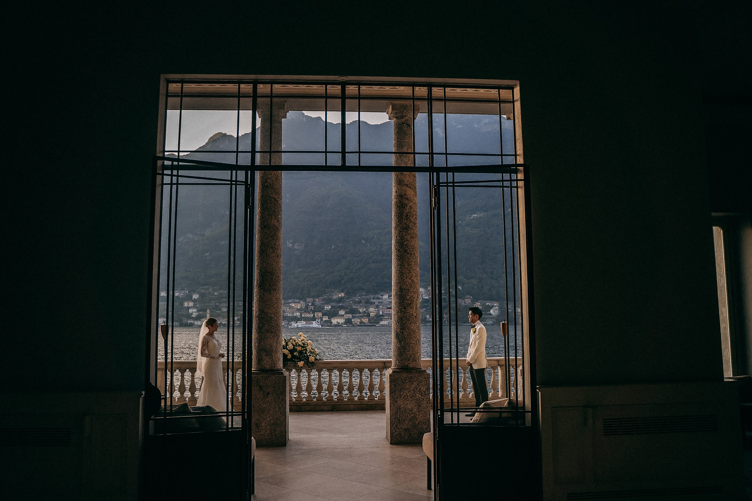Bride and groom face each other from opposite ends of a grand balcony, framed by tall columns and lake Como views.