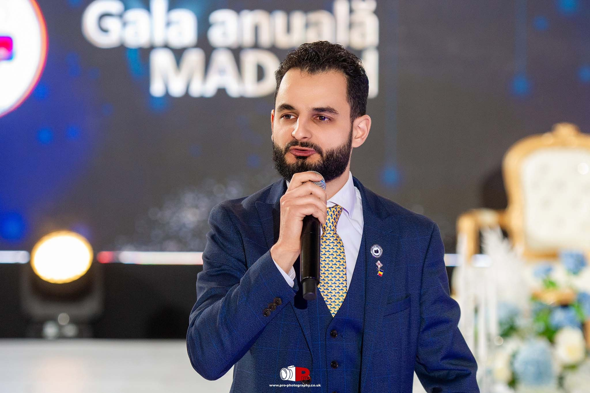 A confident male speaker in a navy suit addressing the audience at the MAD-Aid Gala, with a stage backdrop behind him.