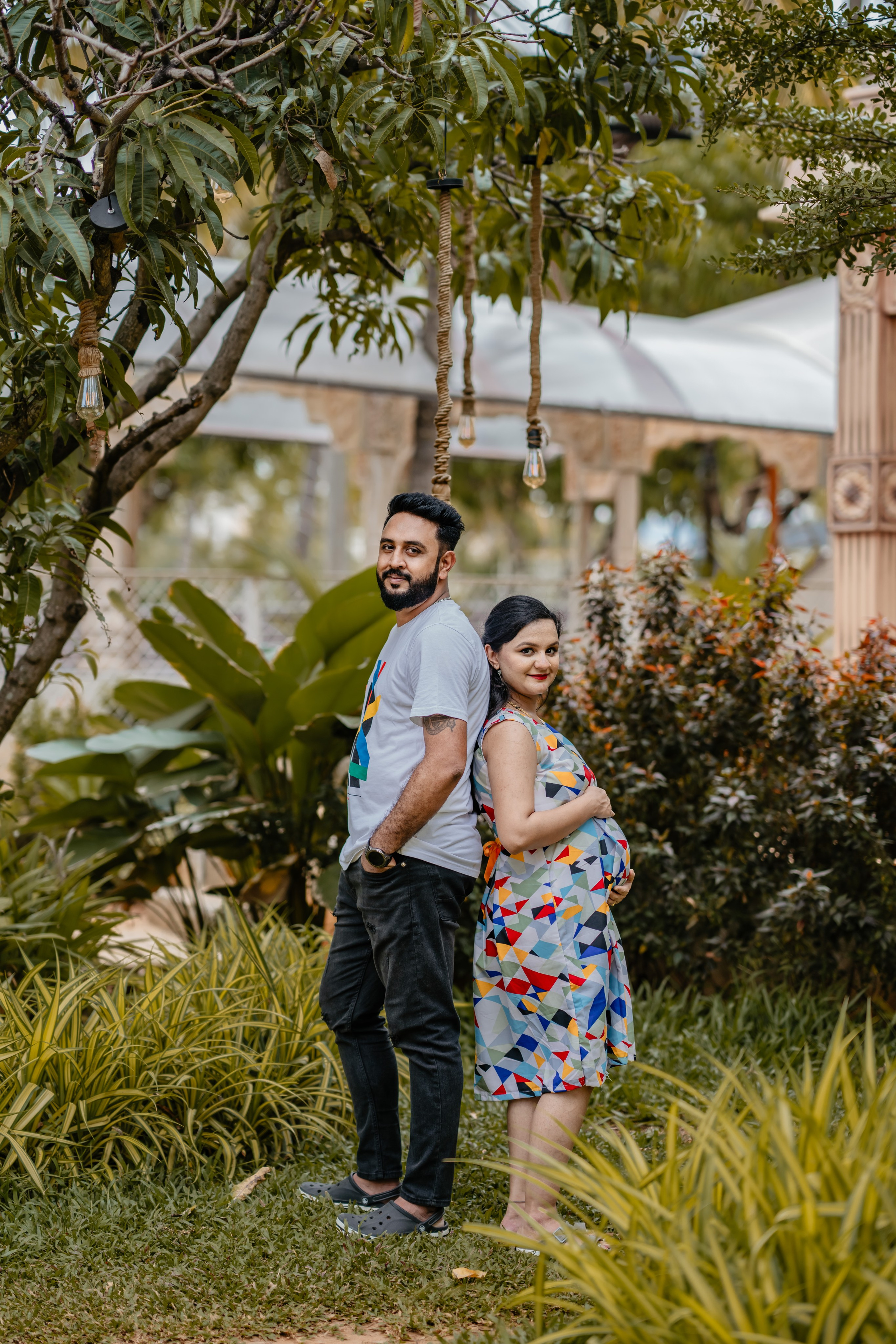 Outdoor maternity photoshoot in Bengaluru featuring a man in a white graphic t-shirt and a woman in a colorful geometric print dress standing back-to-back in a garden studio.