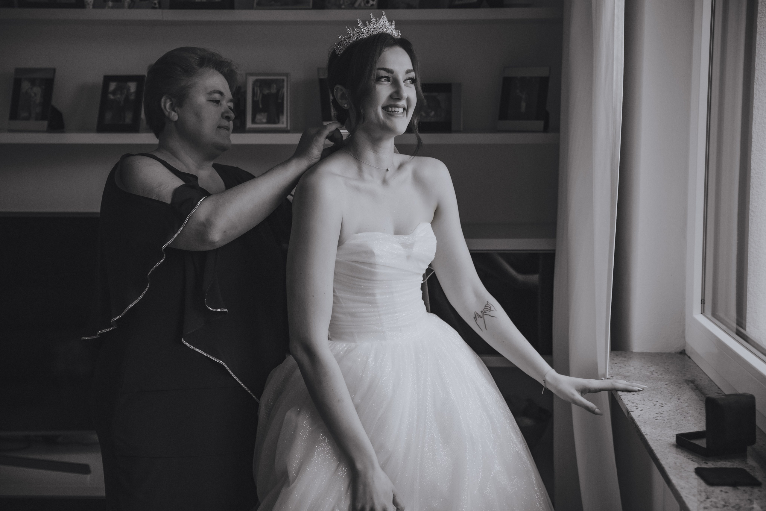 Die Braut schaut aus dem Fenster, während ihre Mutter ihr Schmuckstücke umlegt. Schwarz-Weiß-Foto eines ruhigen, intimen Moments.