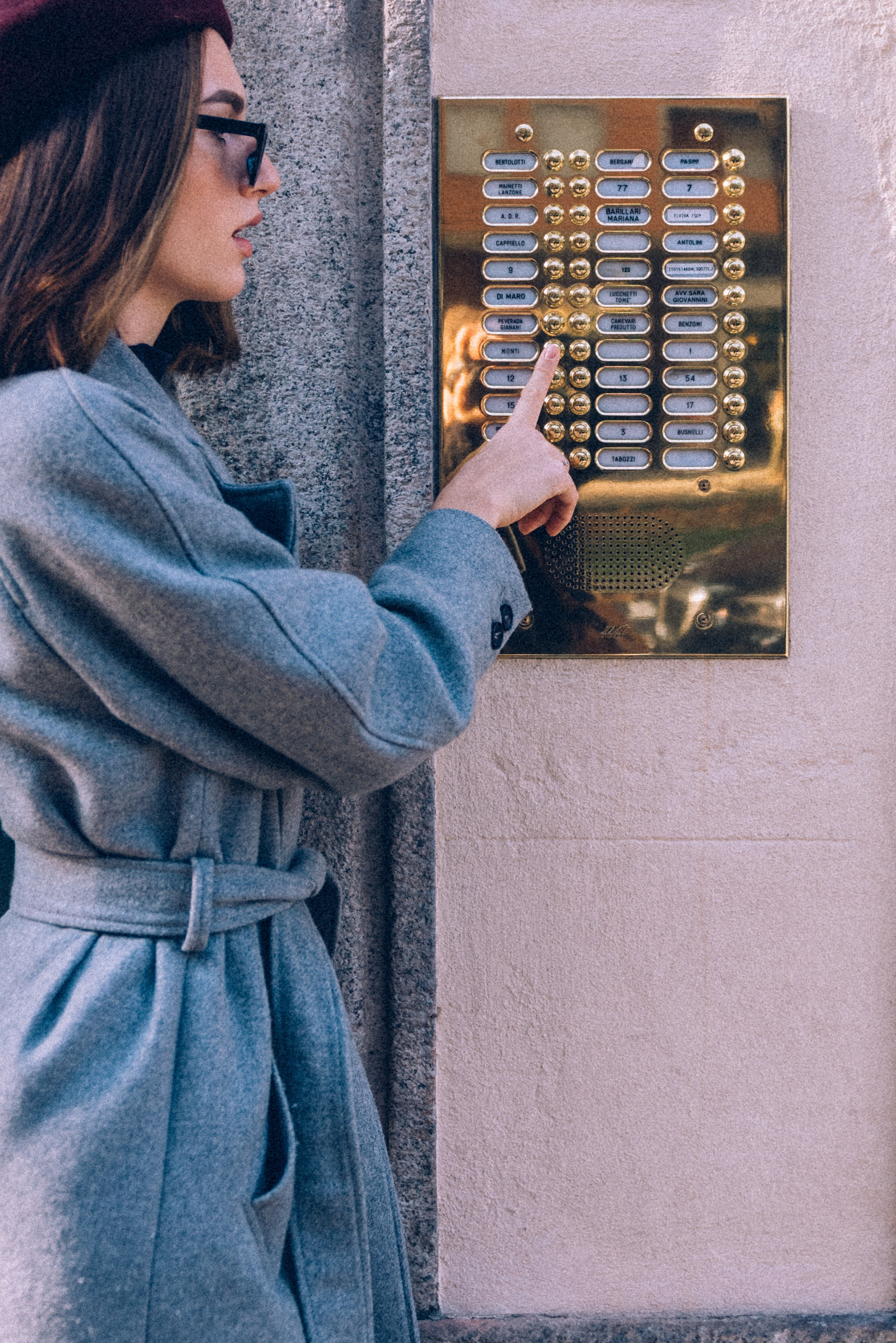 Fashionable woman in a gray coat and burgundy beret interacting with a golden intercom in Milan, capturing urban elegance and everyday life