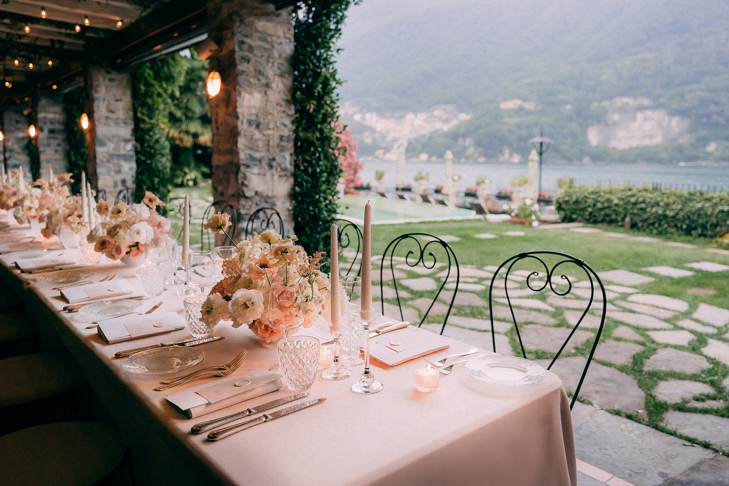 Romantic outdoor wedding reception table under stone patio with lake and mountain views — elegant floral centerpieces, crystal glassware, and candlelit ambiance for luxury event photography.