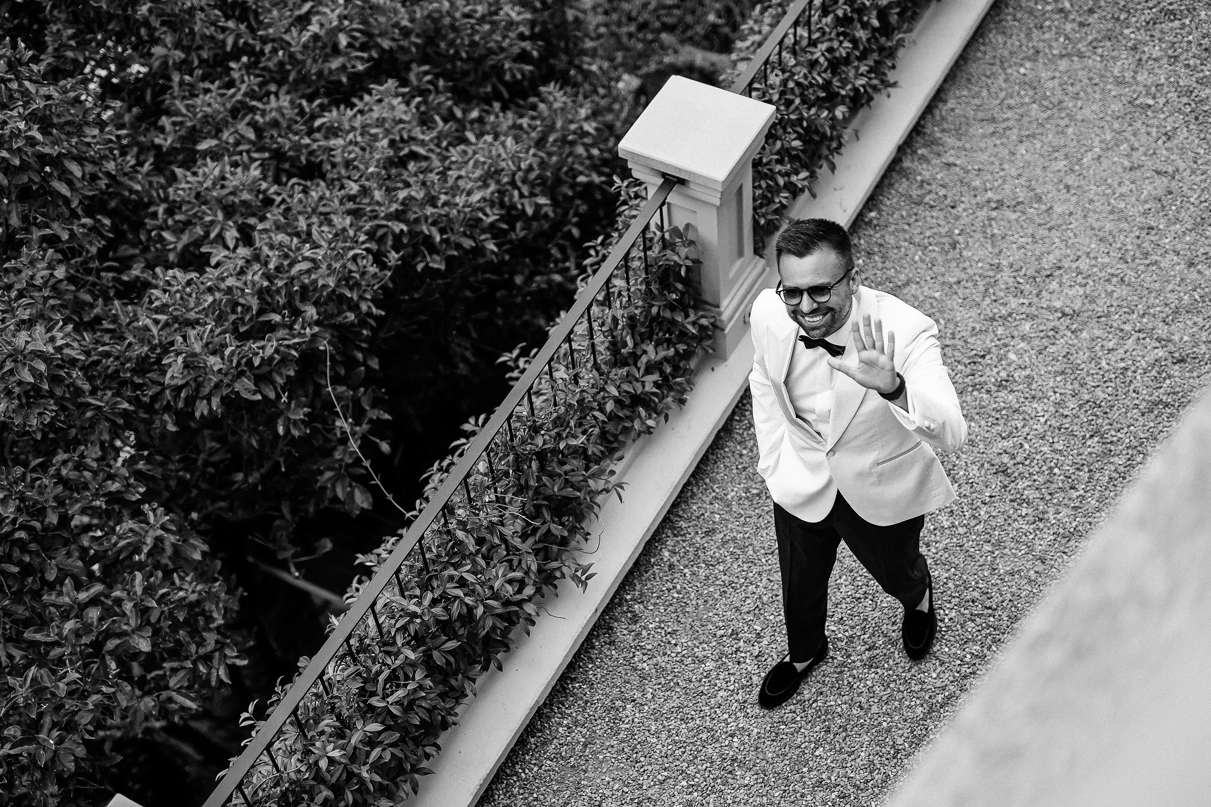 Black and white photo of a groom in a white jacket and dark trousers walking along a gravel path bordered by lush greenery and a railing. The groom gestures with one hand, creating a candid, expressive moment viewed from above.