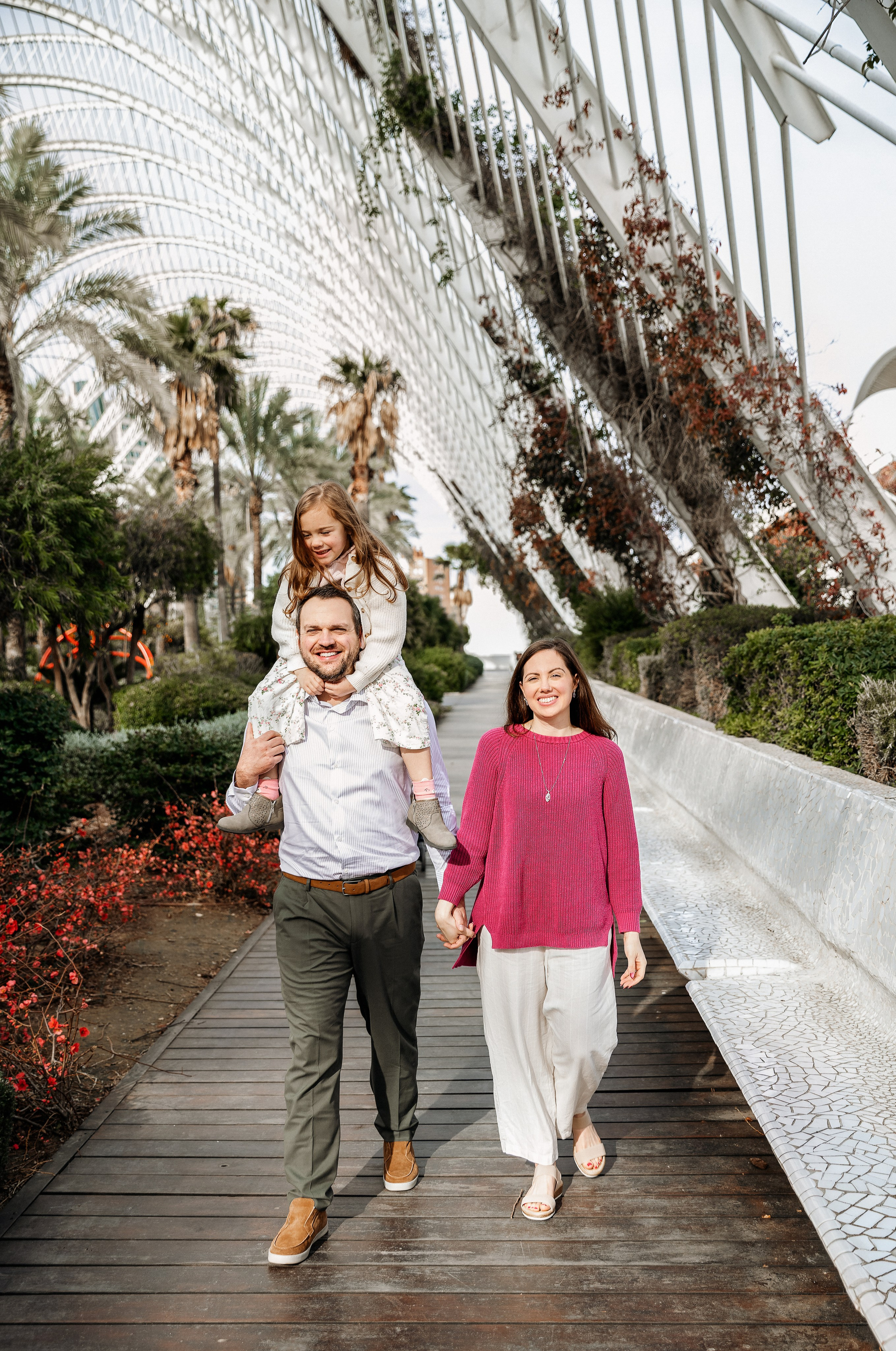 Семейная фотосессия в Валенсии, Испания, где родители гуляют вместе по дорожке, окружённой пальмами, в светлой уличной локации, снято при естественном свете для расслабленного и живого семейного момента — идеально для семейных фотосессий в Валенсии и по всей Испании.