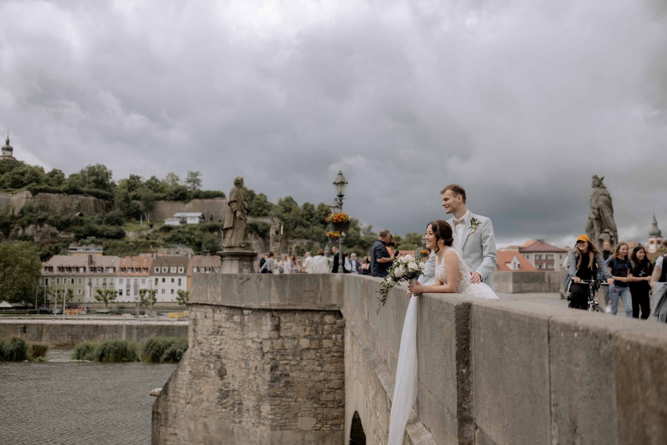 Die Braut blickt über die Brüstung der Brücke in Richtung Festung Marienberg – ihr Schleier und Kleid wehen leicht im Wind, eine friedliche, träumerische Stimmung.