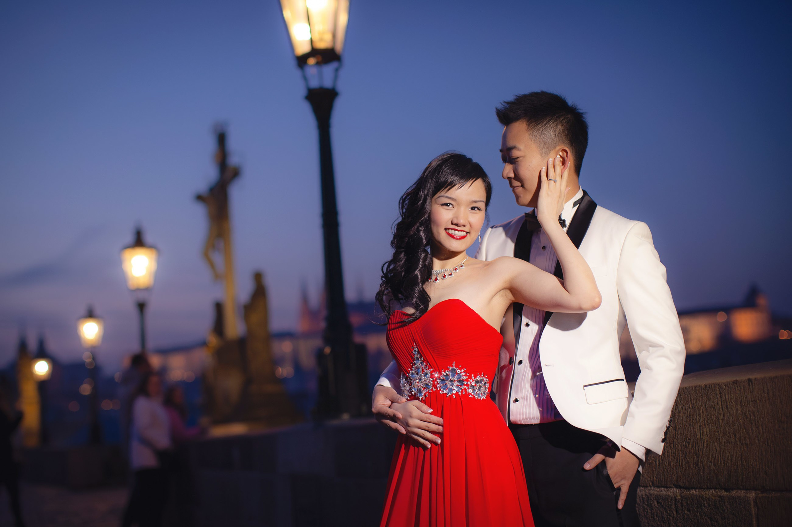 Joyful asian woman in red evening dress is lovingly embraced from behind by her partner dressed in a white tuxedo atop the night time scenery of the Charles Bridge