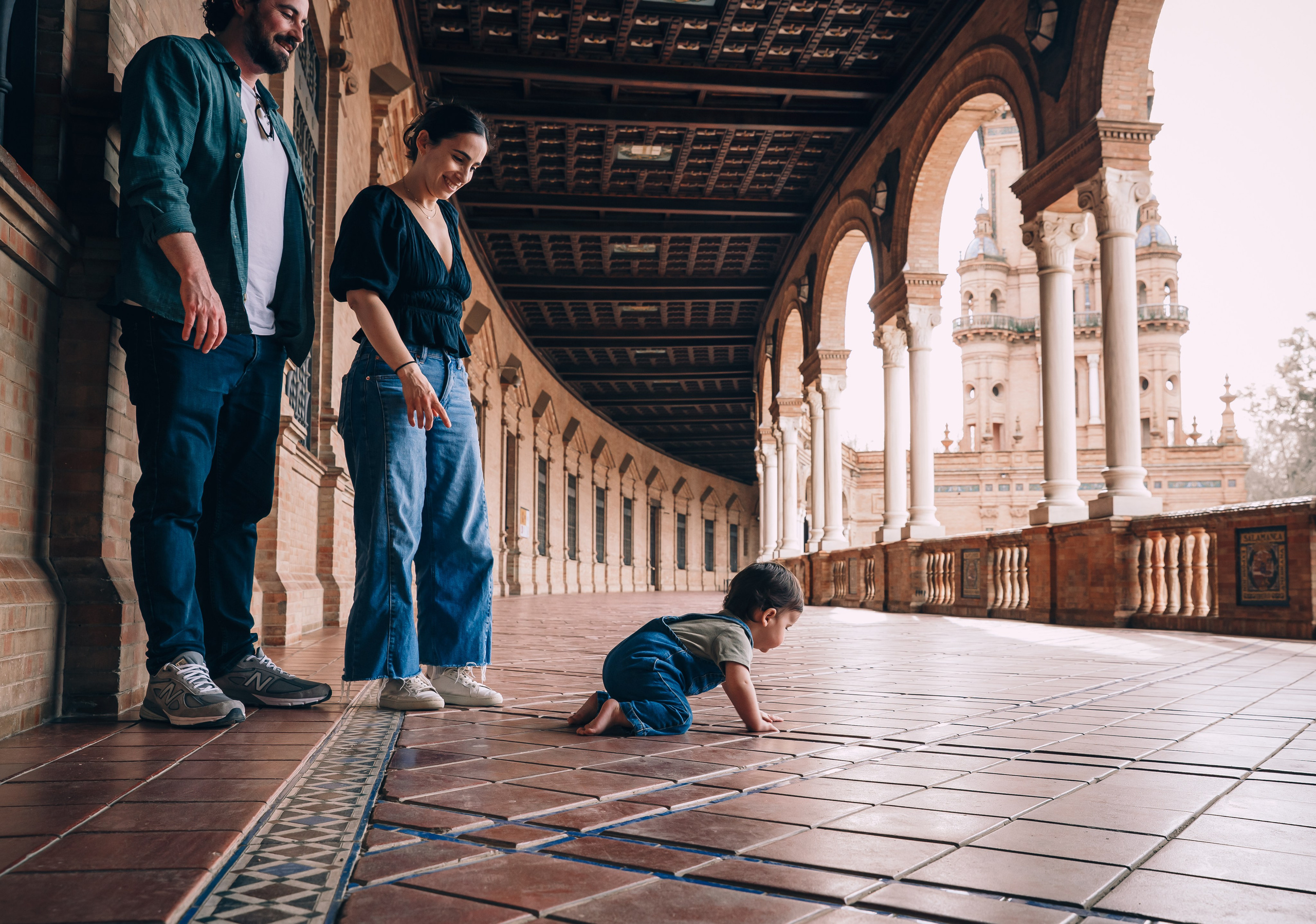 Escena familiar espontánea en la Plaza de España de Sevilla — padres observando a su bebé gatear sobre los azulejos bajo los grandes arcos, rodeados de la majestuosa arquitectura española.