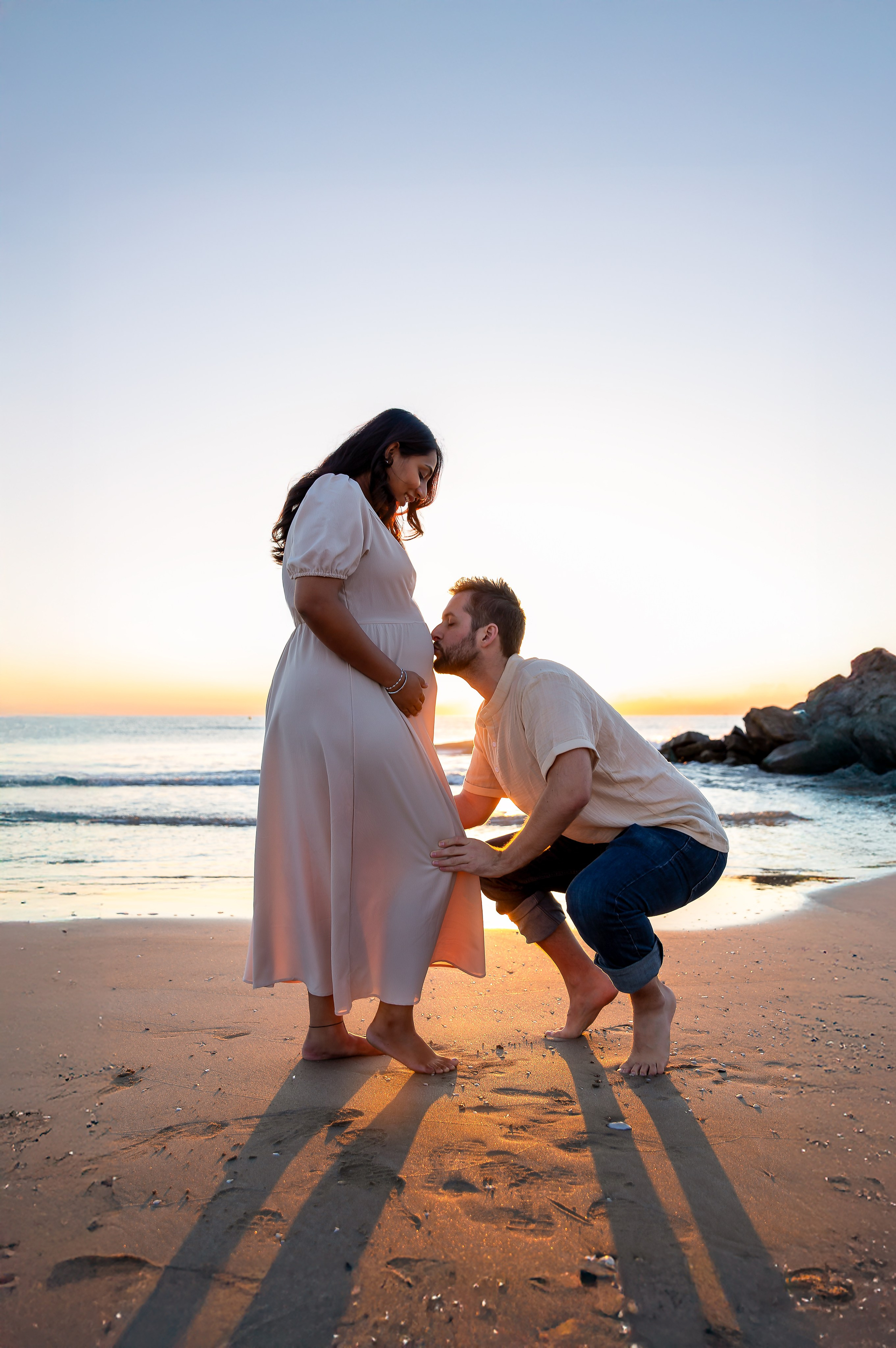 Dulce sesión de maternidad en la playa de Valencia, España — futuro padre arrodillado besando la barriga de su pareja embarazada al atardecer, capturando un instante romántico y lleno de emoción. Inspiración ideal para sesiones de embarazo en Valencia y en zonas costeras de España.
