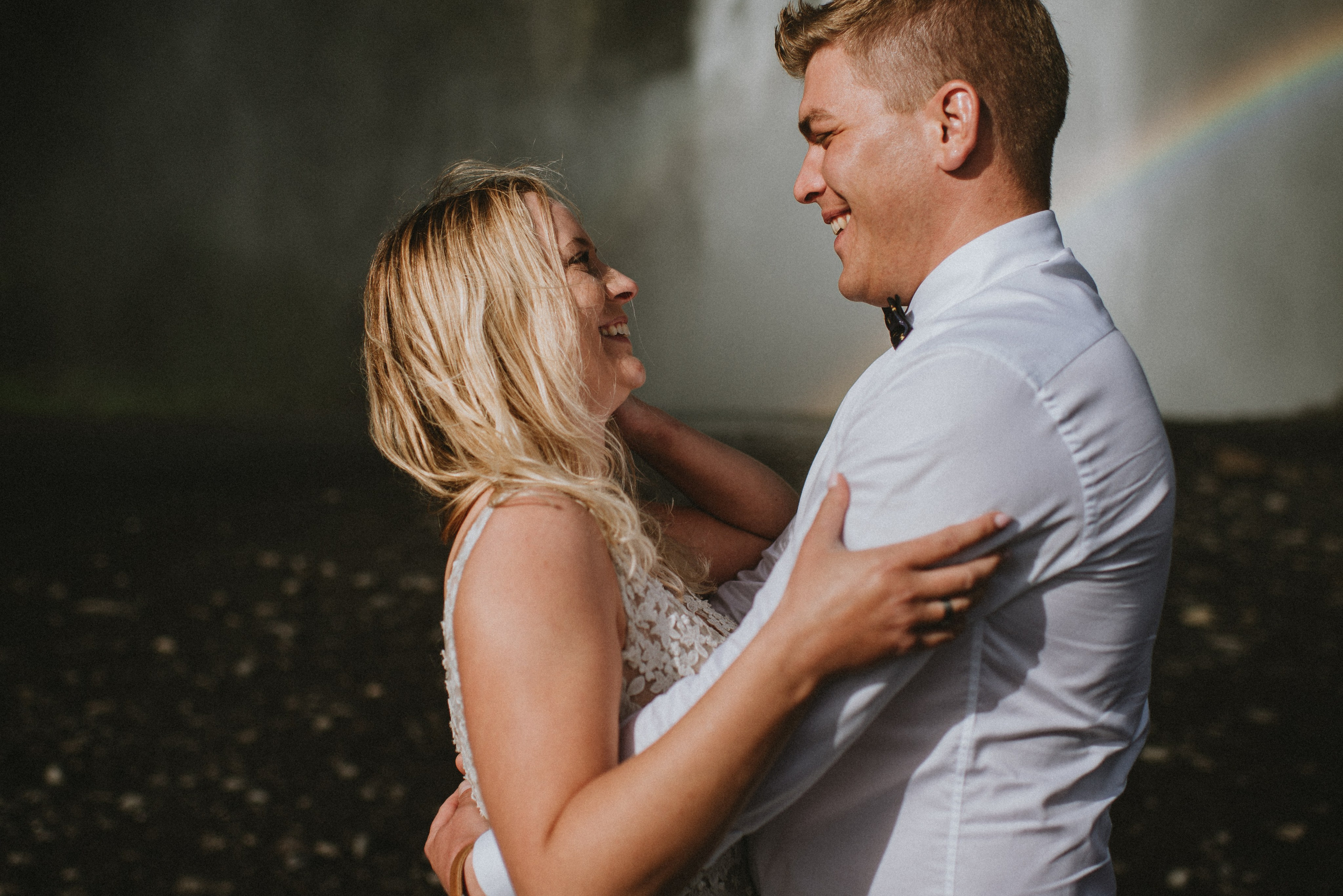 Bride and groom looking up at the towering Skógafoss waterfall, holding hands.