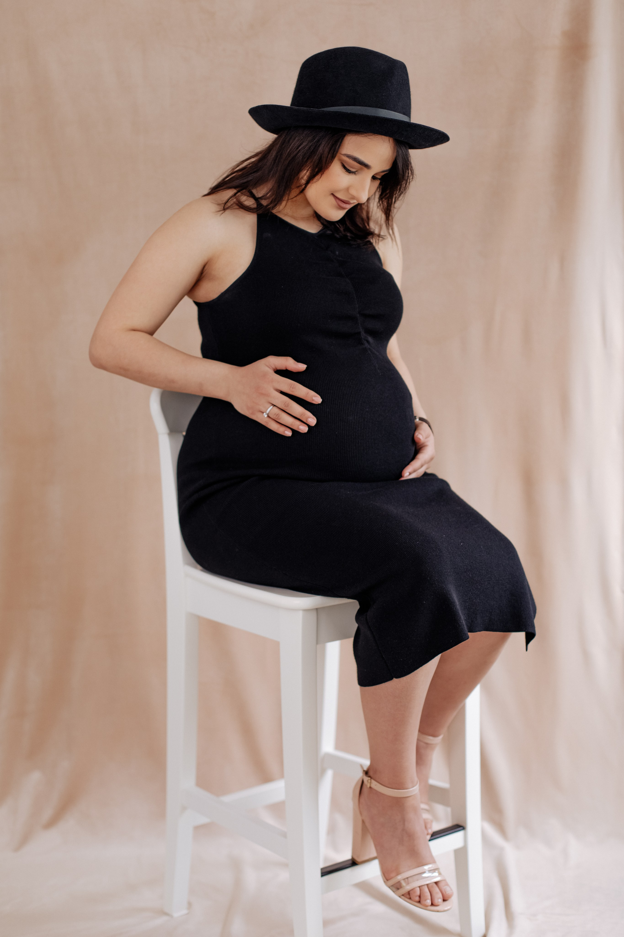 Stilvolle schwangere Frau mit Hut sitzt auf einem hohen Stuhl während eines Schwangerschaftsshootings.