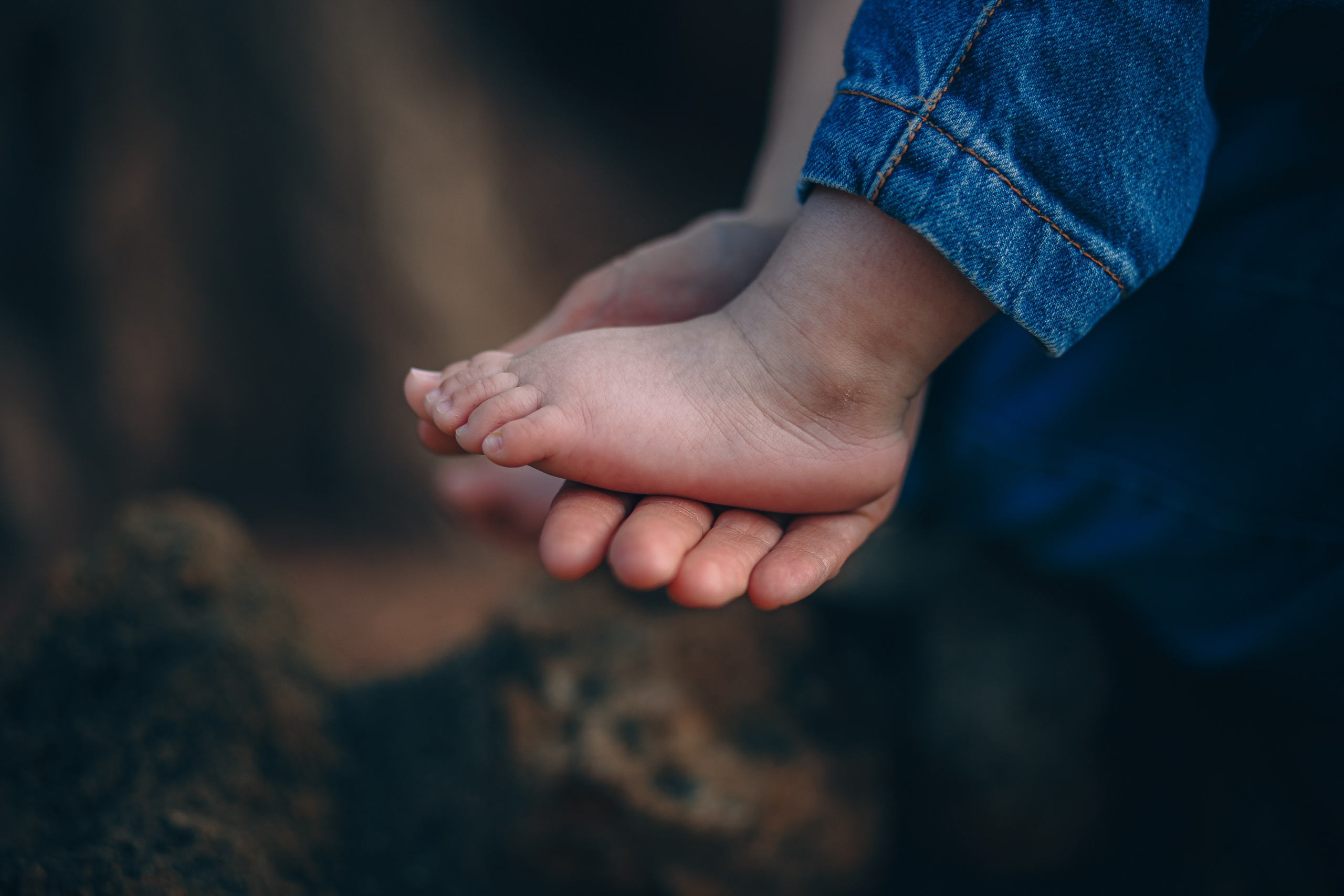 Primer plano de un padre o madre sosteniendo el pequeño pie descalzo de su hijo, con luz natural suave y tonos terrosos de fondo — un momento familiar conmovedor capturado al aire libre en Valencia, España.