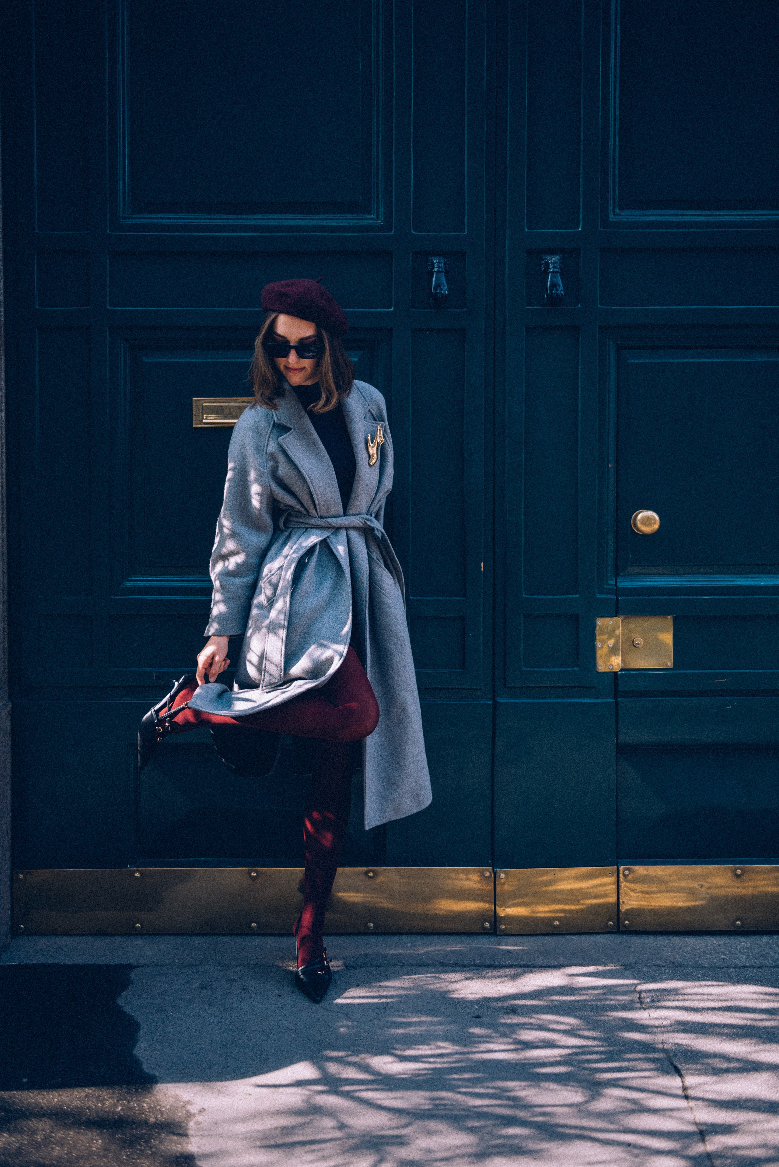 Playful and stylish: a woman strikes a pose against a classic Milanese door, wearing a gray coat and burgundy tights, combining fun with elegance