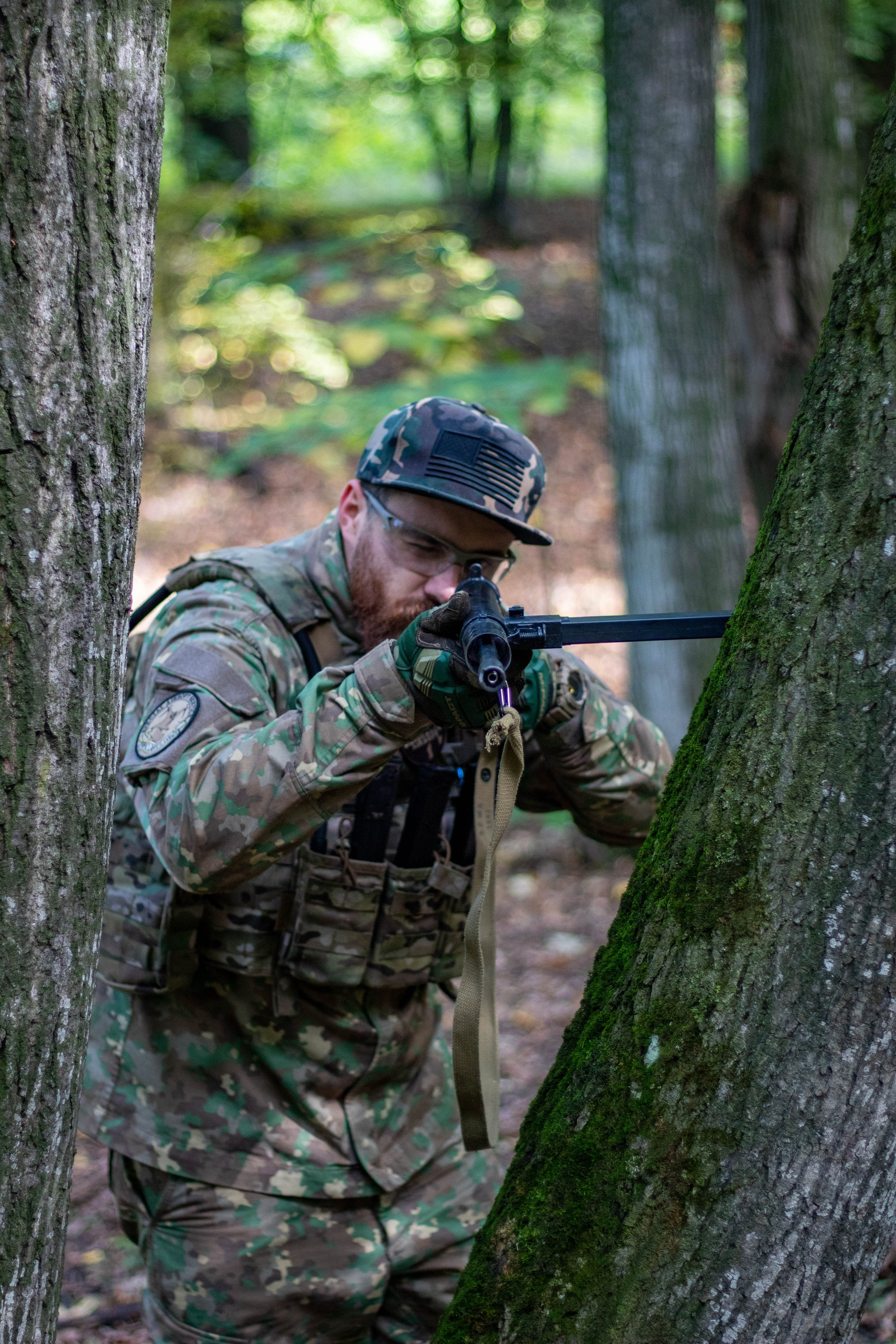 Camouflaged airsoft sniper aiming through a scope from behind a tree in dense woodland.